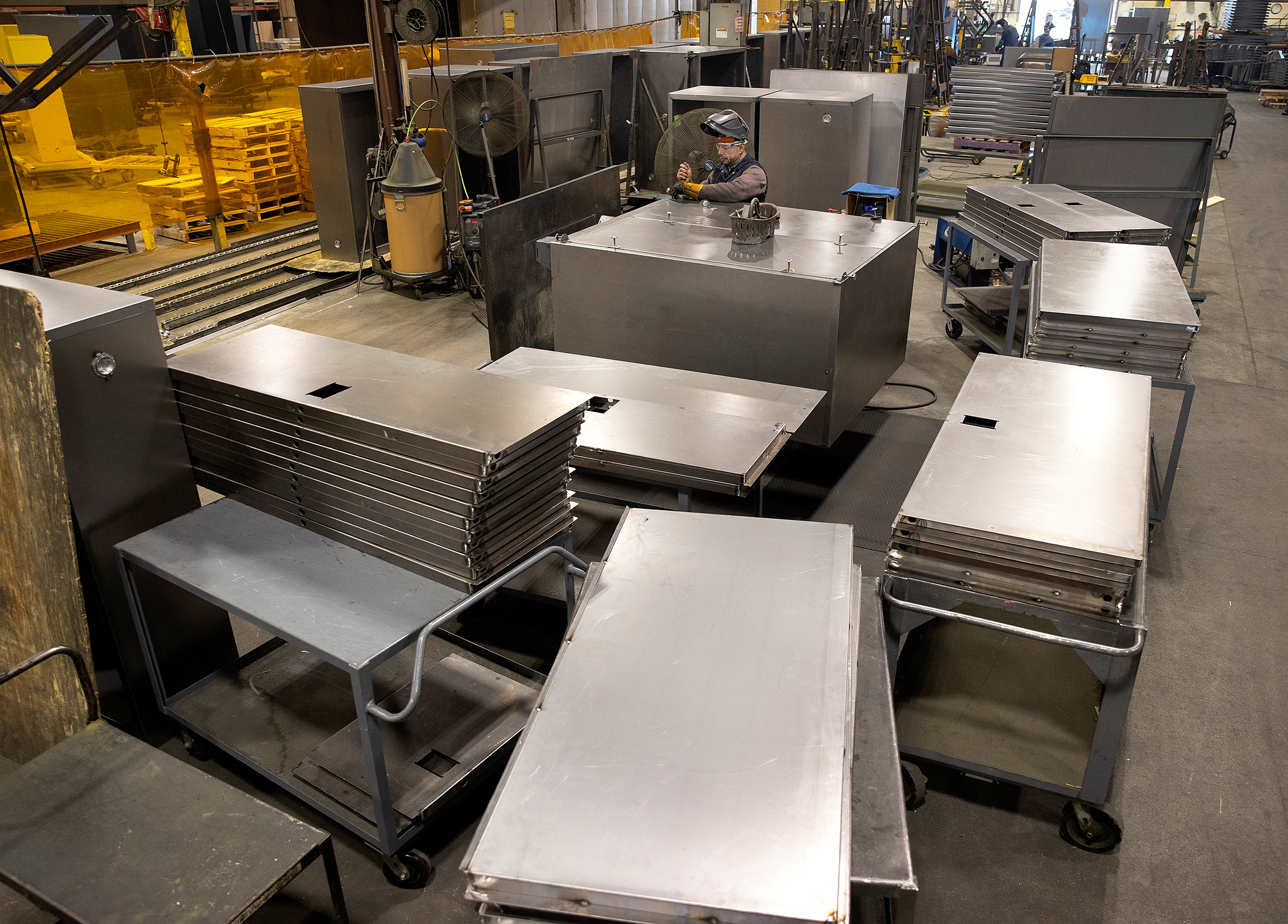 A worker rivets together pieces of metal into cabinets in a manufacturing facility. The worker is in the back of the photo, with stacks of flat stainless steel pieces in the foreground. The worker is wearing protective safety equipment.