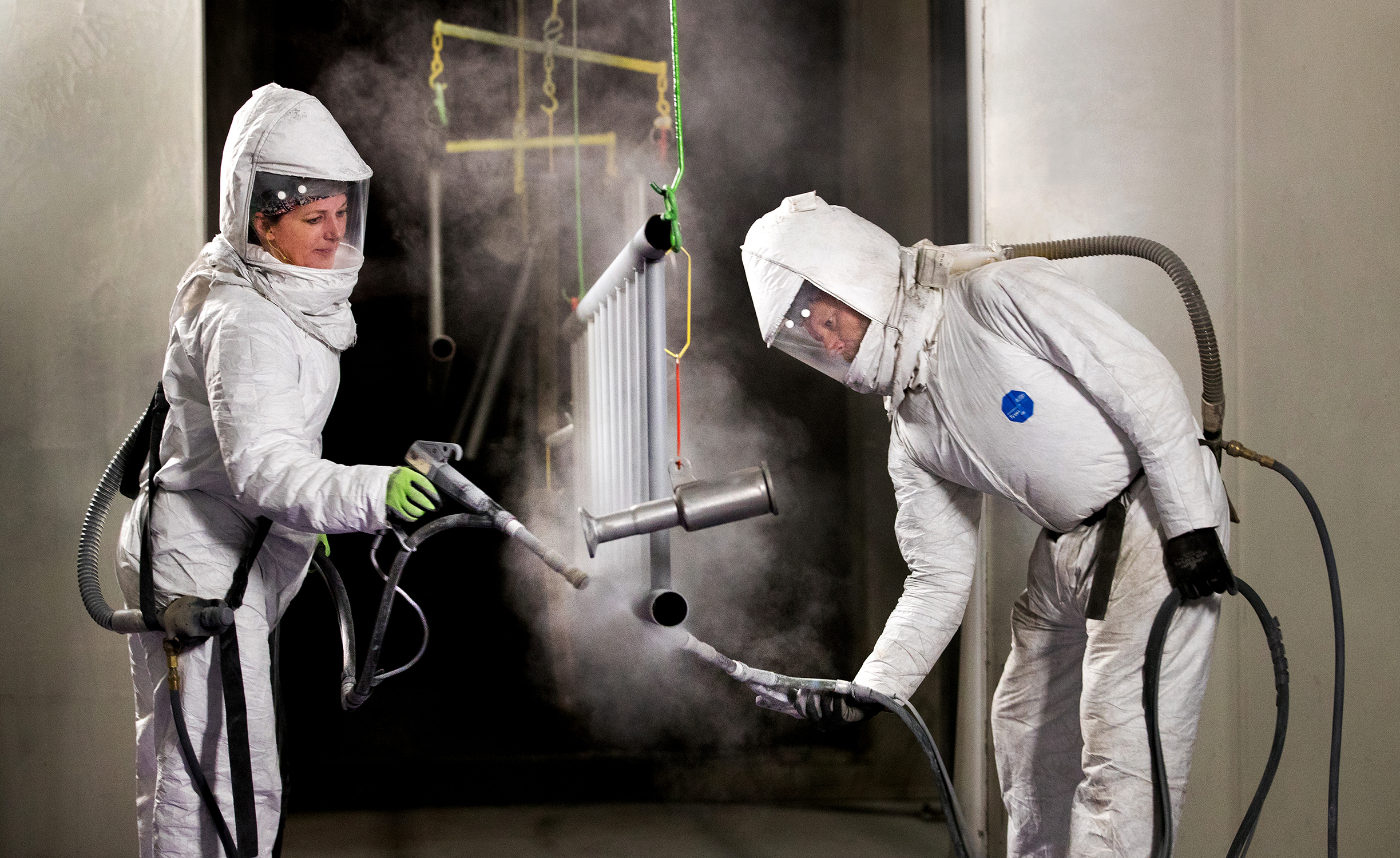 Two workers wearing full-body suits with separate respiration tubes are painting pieces in a manufacturing facility. Both workers, who are women, are spraying paint onto the components, which are hanging in the center of the image. The workers are hard at work.