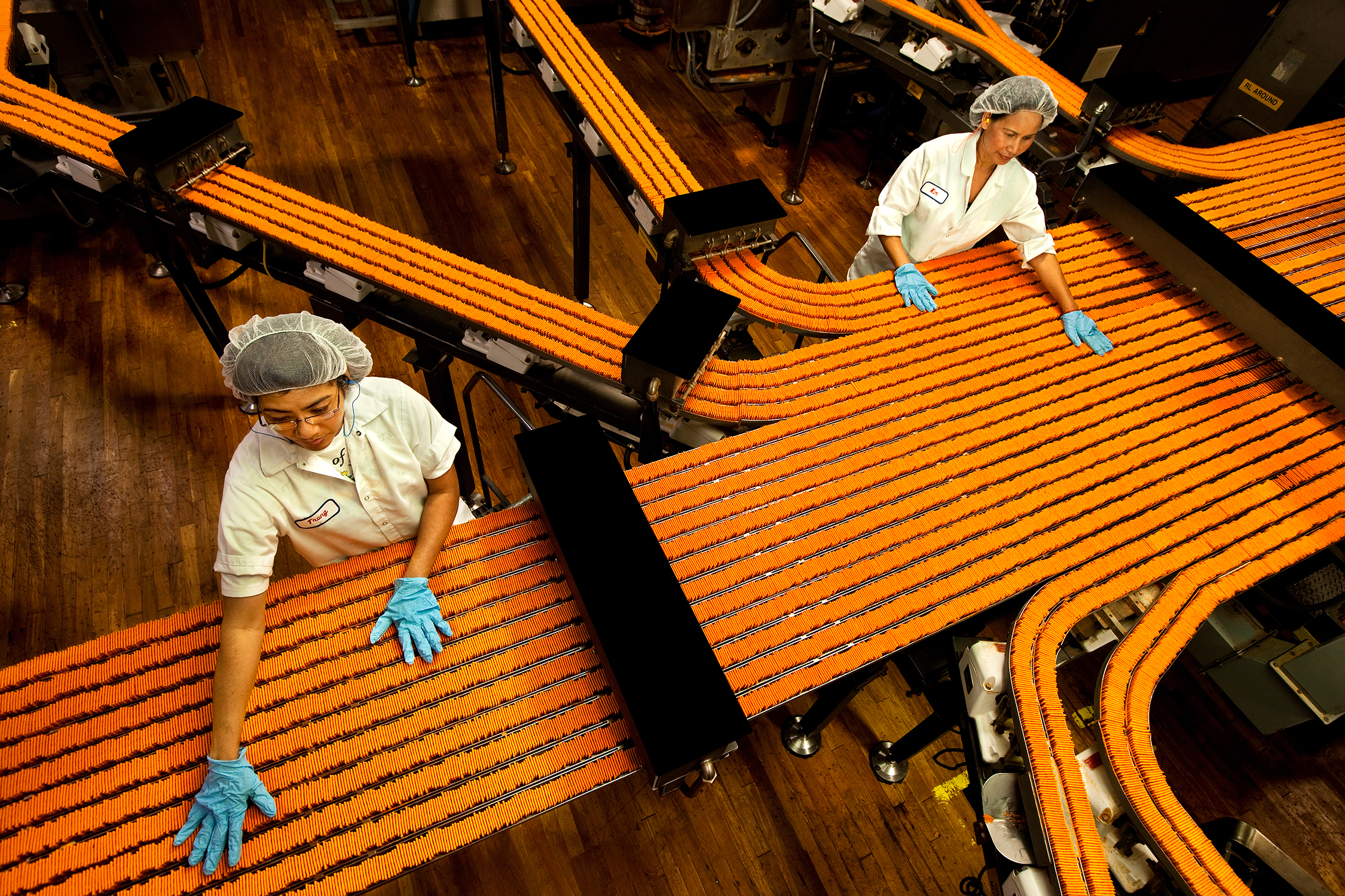 Two women watch the lines of cheese crackers flowing through a food processing and manufacturing facility. The women are standing with their gloved hands on the crackers, which are flowing by them. Both women are wearing proper hygiene attire, including coats, gloves, and hairnets.