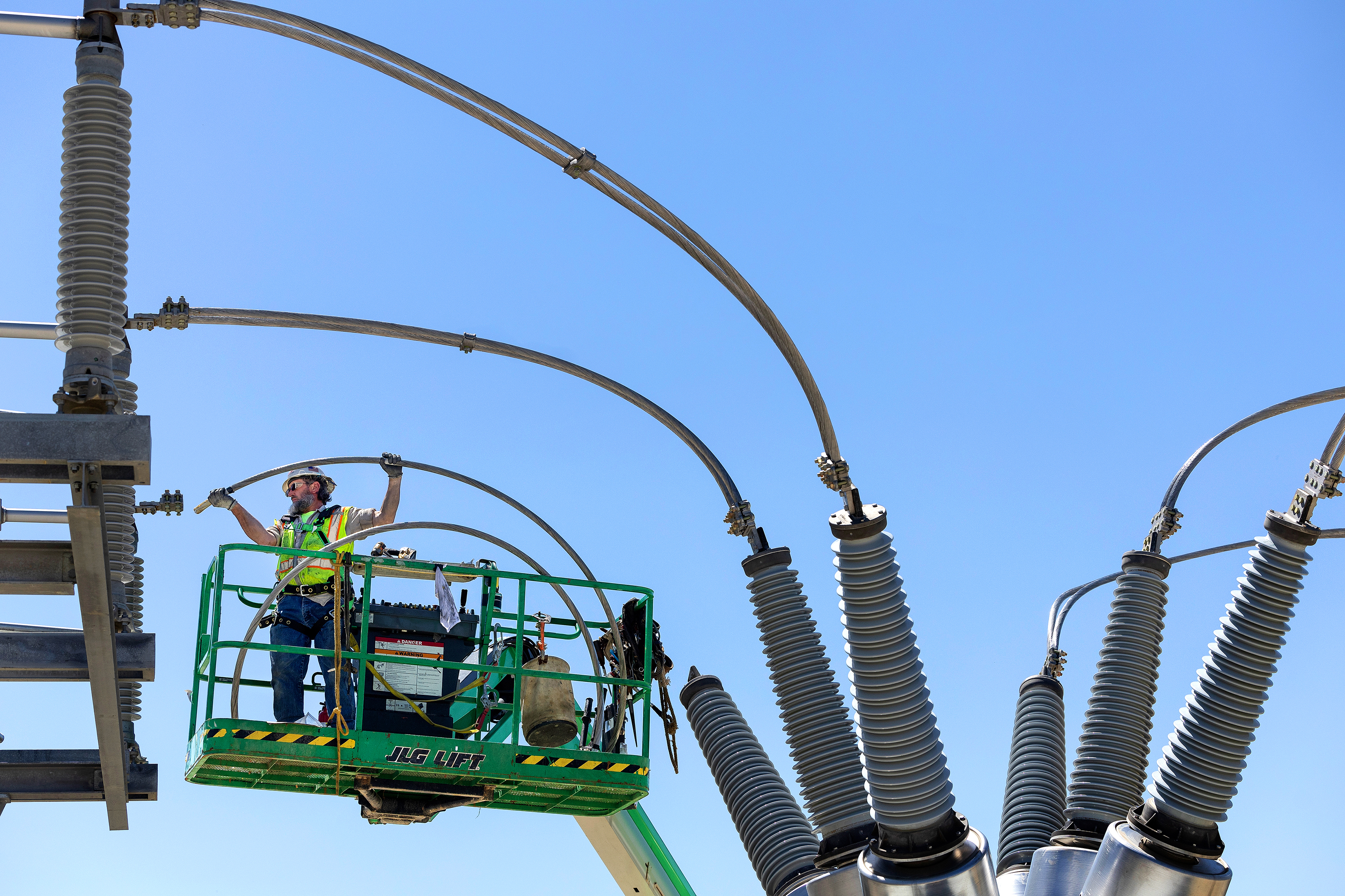 Photograph of a male utility worker connecting giant cables. The worker is wearing proper protective gear and is standing on a lift. Image was taken using a drone and is on the same level as the worker.