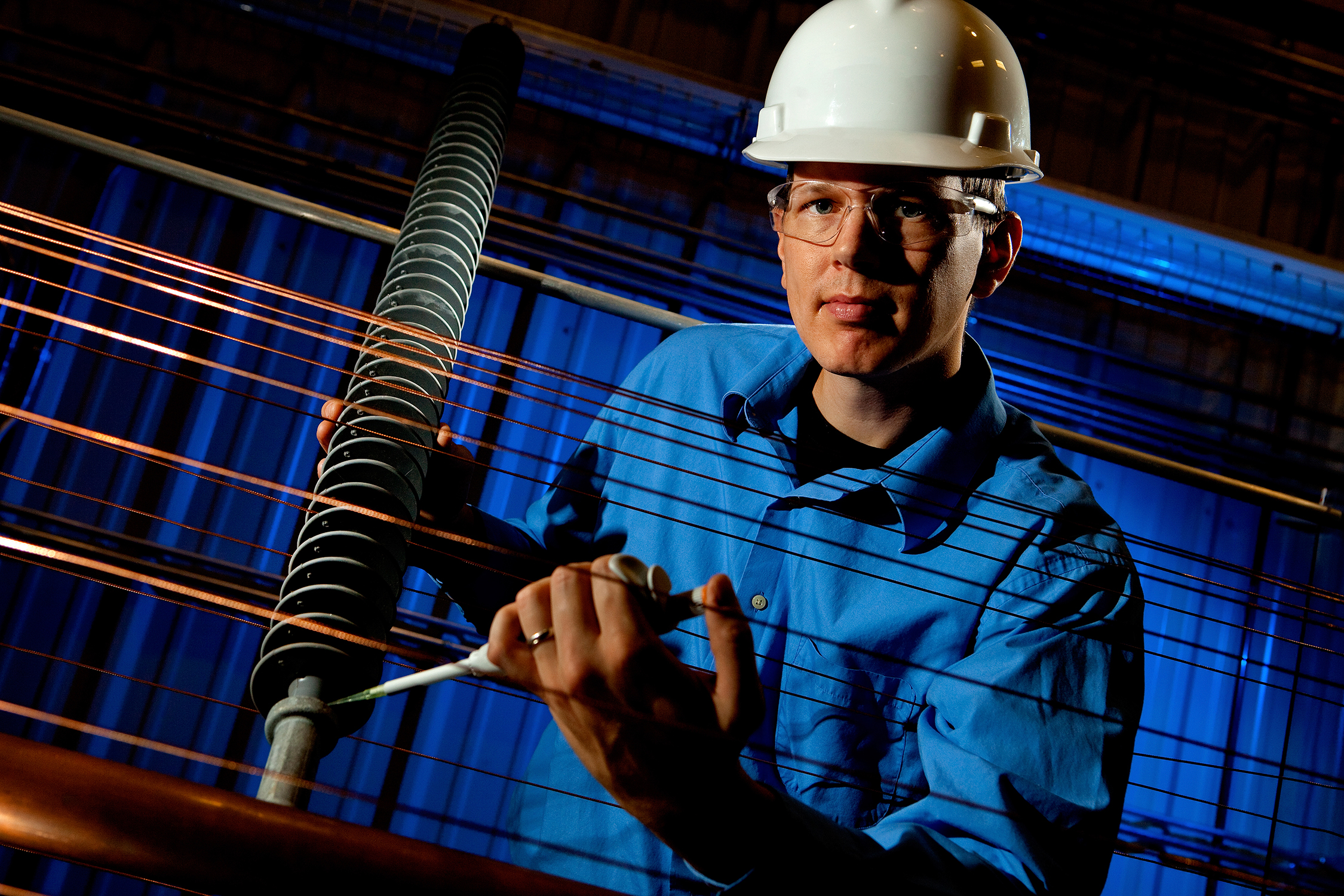 Photo is an environmental portrait of a male worker on a high tech assembly line. The man, who is wearing a hardhat and safety glasses, is looking at the camera while he is adjusting the manufacturing line.