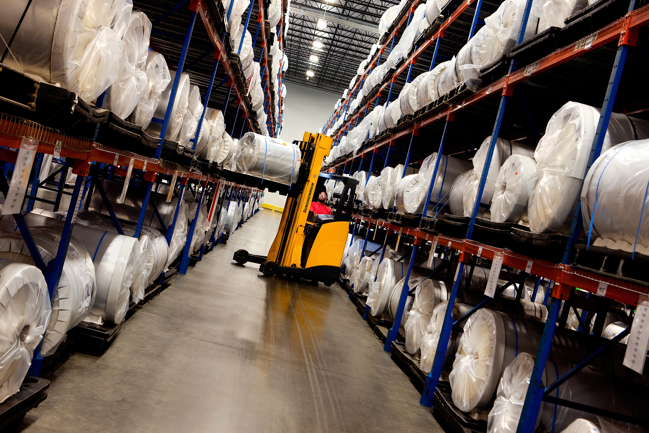 A forklift raises a large role of material onto the shelves of a manufacturing plant. Hundreds of rolls of similar materials rest on the shelves, which are at least four levels high. The photo is taken so that the forklift driver is visible and can be seen working in the background of the image.