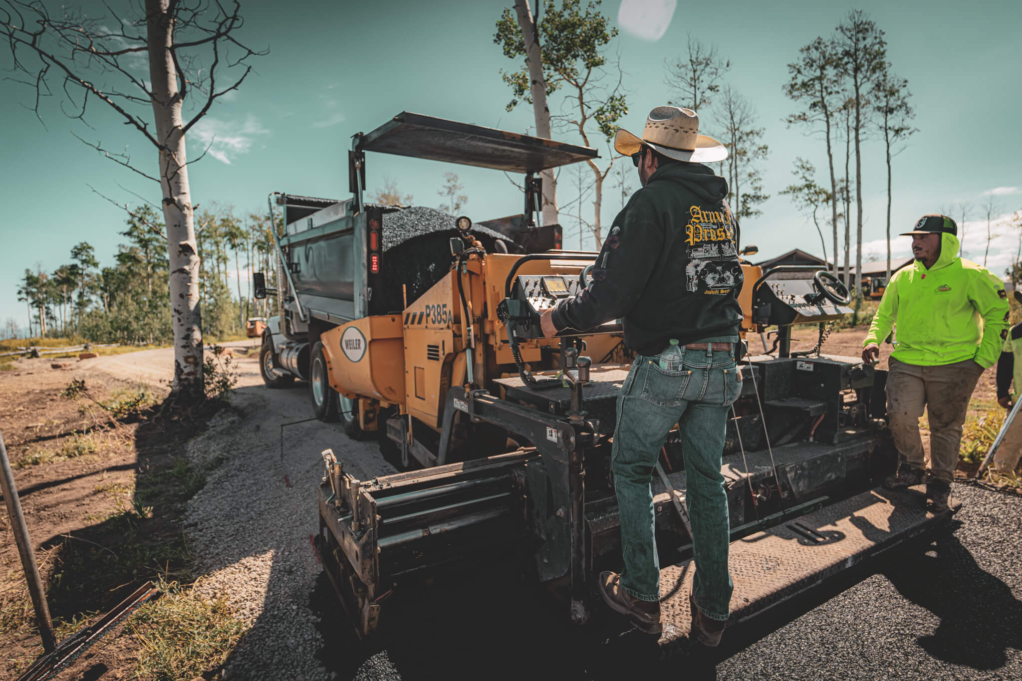Armor Proseal crew installing new asphalt pavement in Pagosa Springs, CO, with professional paving equipment and workers in safety gear operating in a wooded residential area.