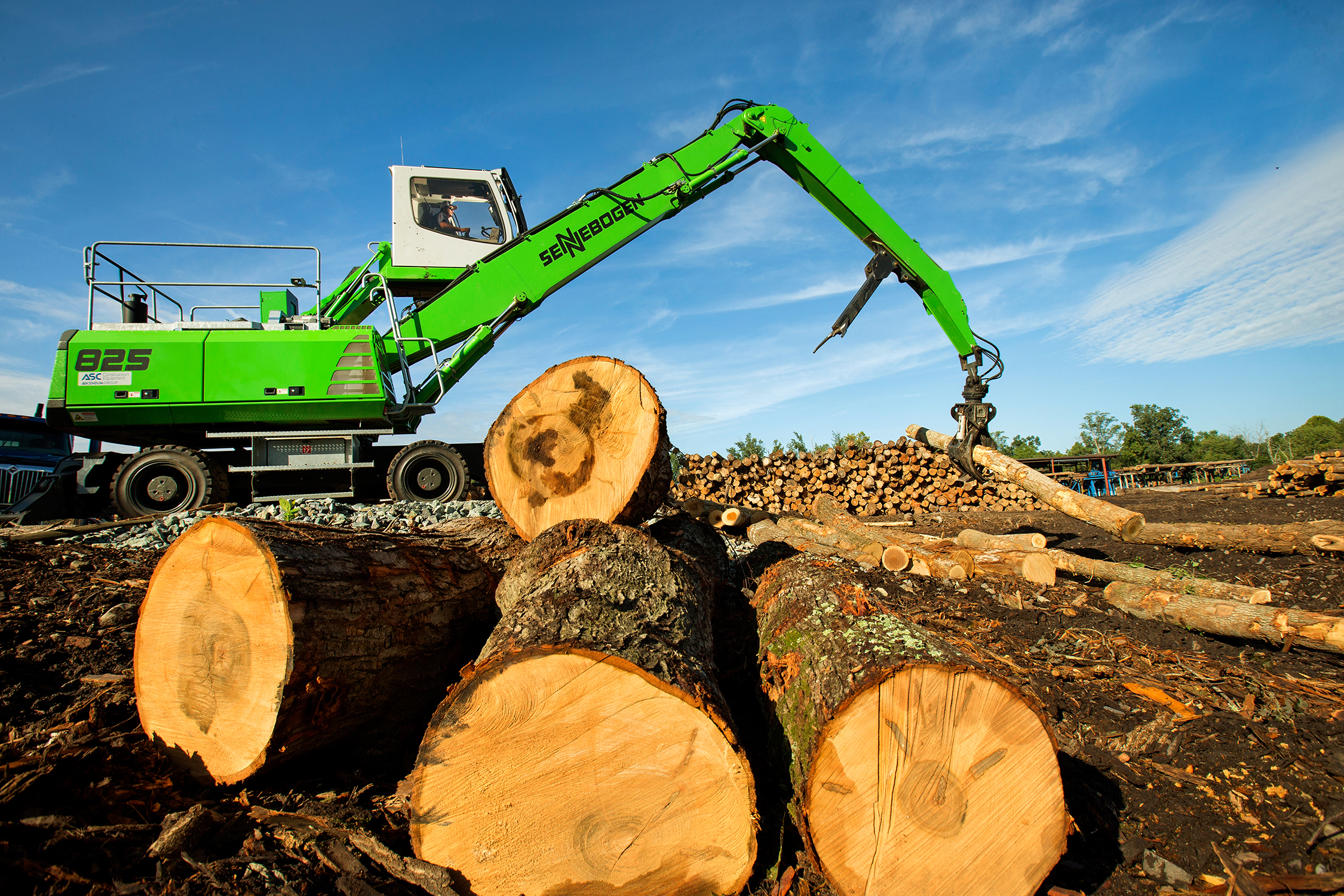 The driver of machine with a giant arm lifts large logs onto a pile of logs at a mill. The cab of the machine is lit with strobe lighting to make it possible to see the man operating the machinery. The machine is bright neon green and stands out against the brown stacks of logs, which range in diameter from several inches to several feet.