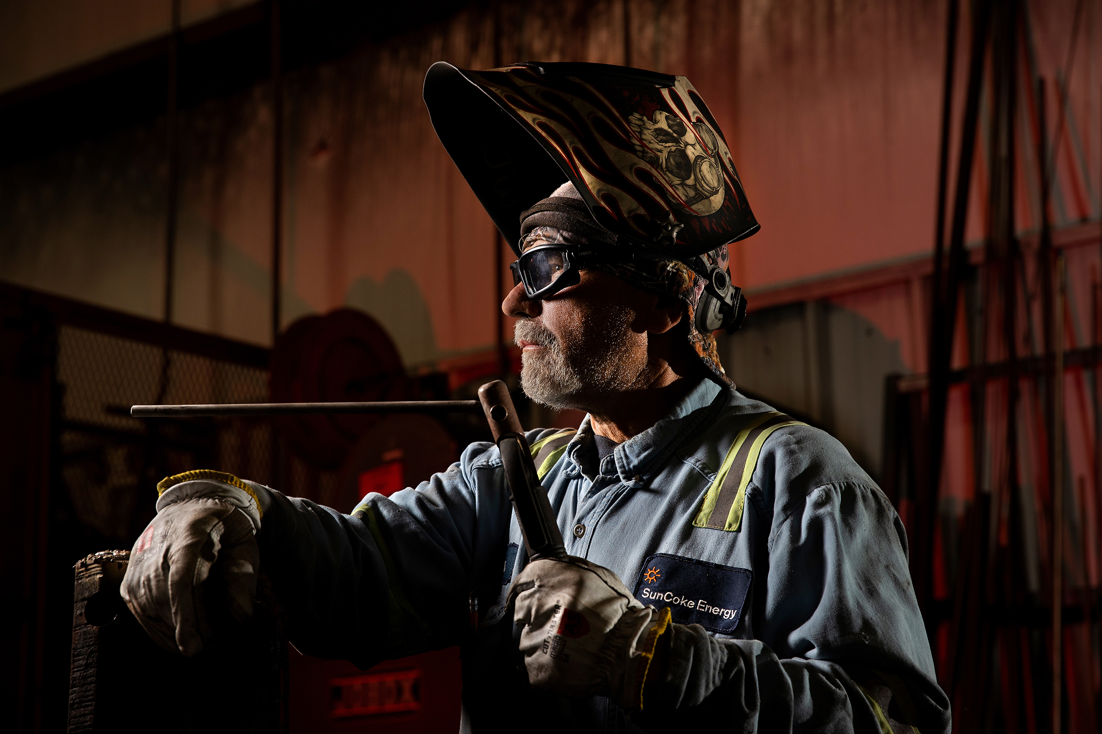 A male worker poses for a photo with his welding hat pushed up so his face can be seen. He is looking off the photo to the left. He is holding a tool in his hand, as if he was just asked to stop working so his photo could be taken. The man is wearing proper safety equipment, including the welding mask, safety glasses, and heavy gloves.