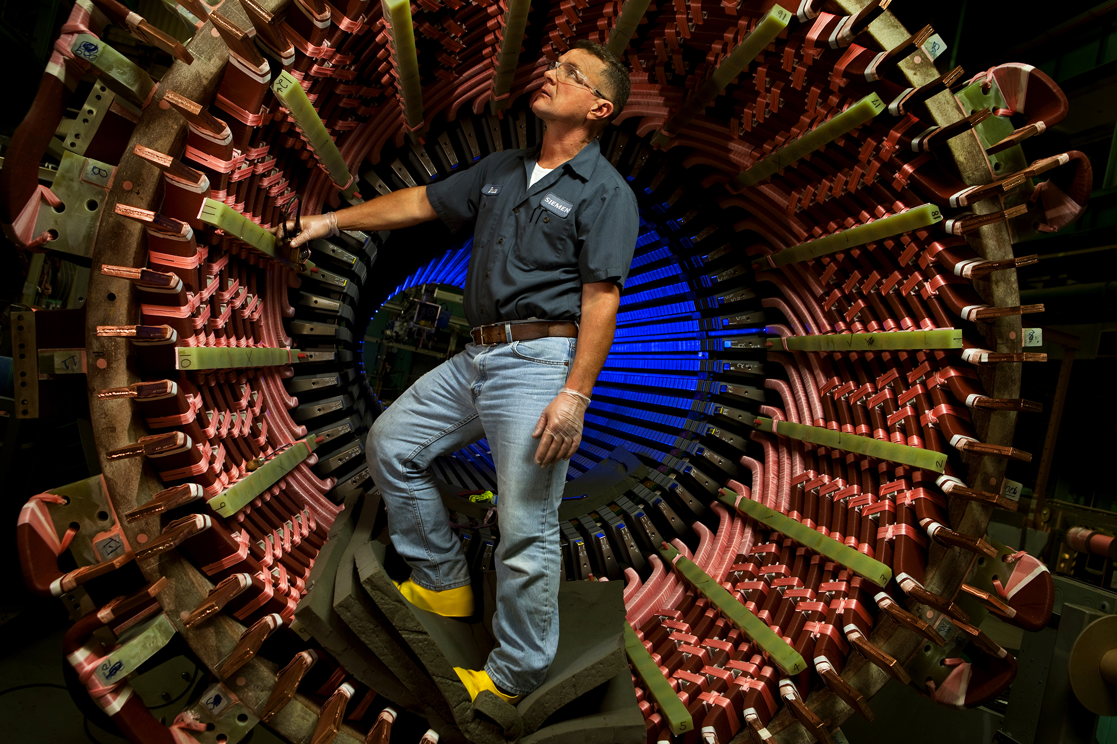 Photo is of a worker inside of an aircraft engine, while the engine is in the manufacturing facility. The engine is taller than the worker, and he is standing inside of the engine. He appears to be inspecting the engine. The worker is wearing safety equipment.