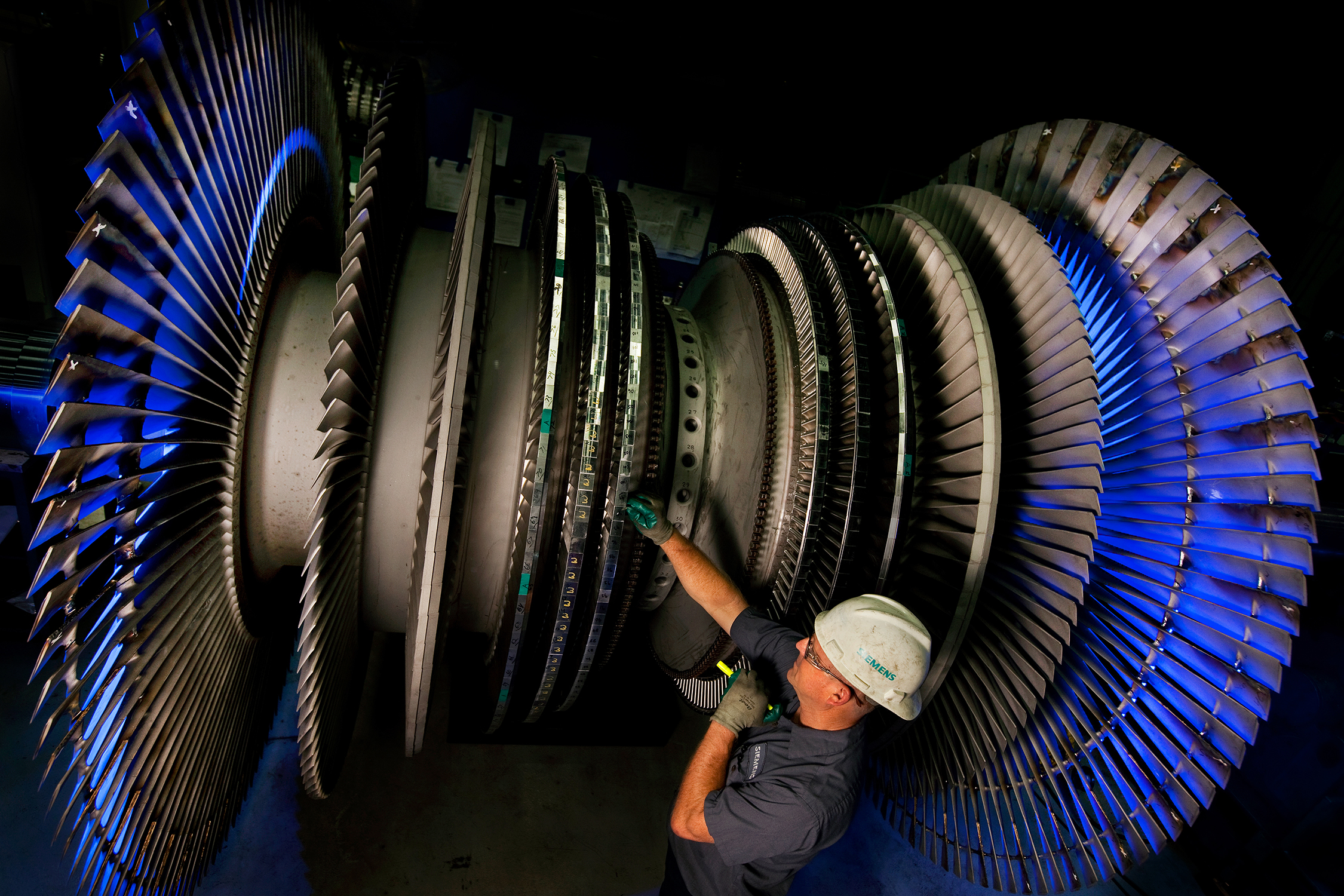 An employee closely inspects the components of an aerospace turbine. He is wearing safety equipment and is using a flashlight to inspect the turbine. The turbine is much larger than the man.