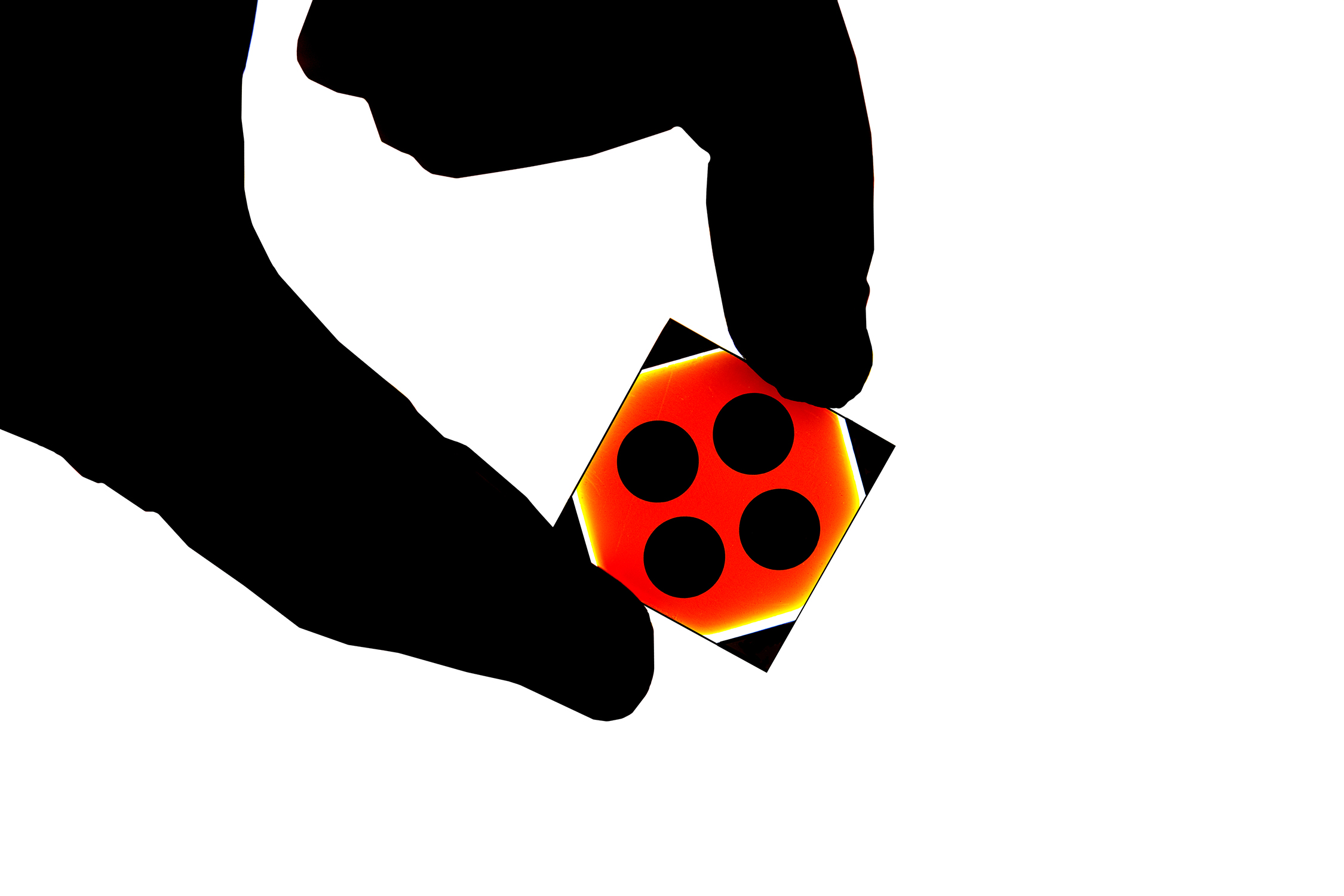 Photo show hand holding a semiconductor chip. The photo is taken close to the hand, as just the thumb and index finger are visible. The chip is red, yellow and black and is very colorful against the silhouetted fingers. The photo was taken in an advanced manufacturing facility or clean room.