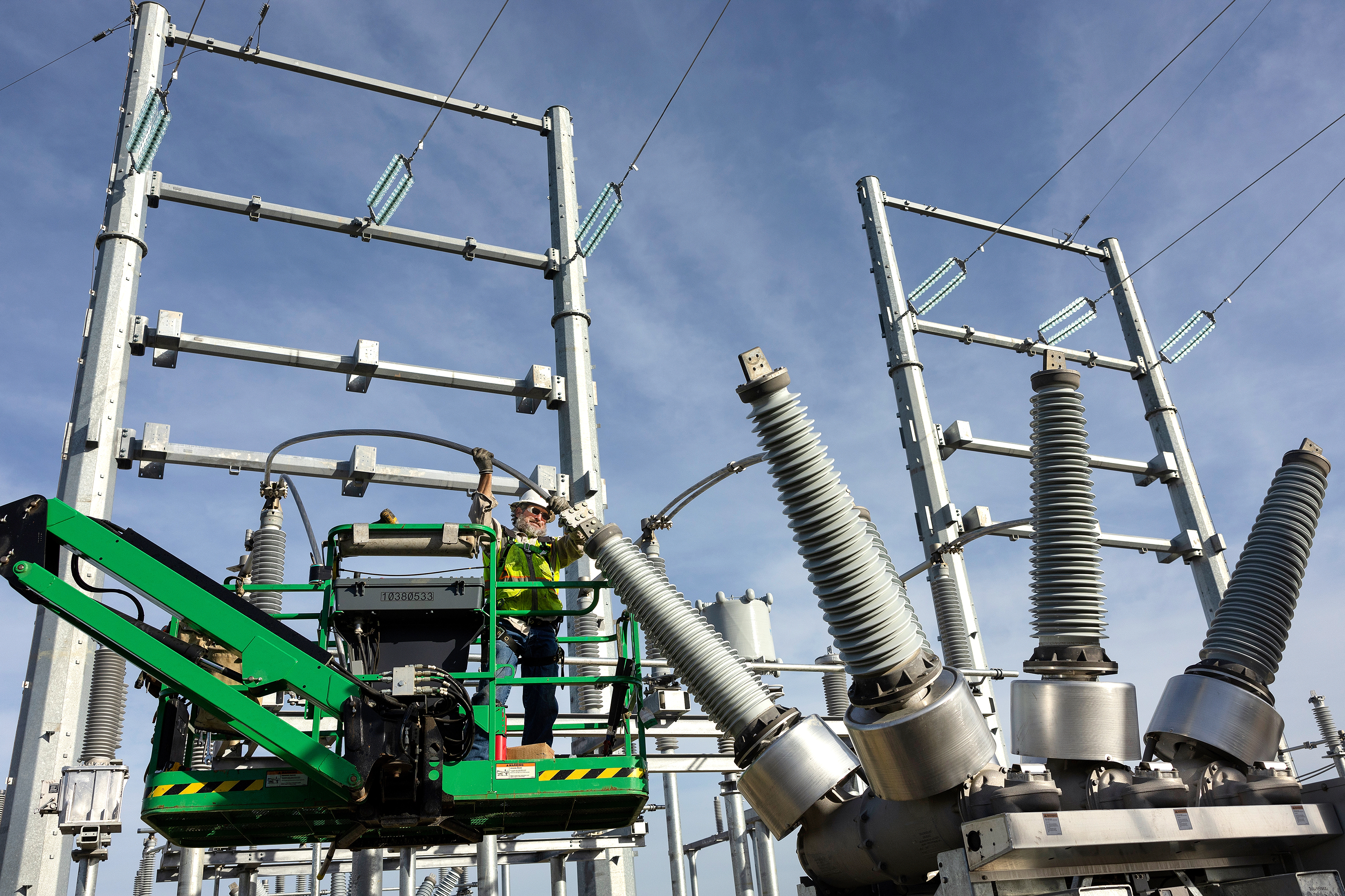 Photo of a utility worker setting up a new electrical facility. He is standing on a lift and is inserting transmission wires into what appears to be a conductor. The photo is taken at the same level of the worker, so the image is straight on. The photographer used a drone to create this image of the electrical worker.