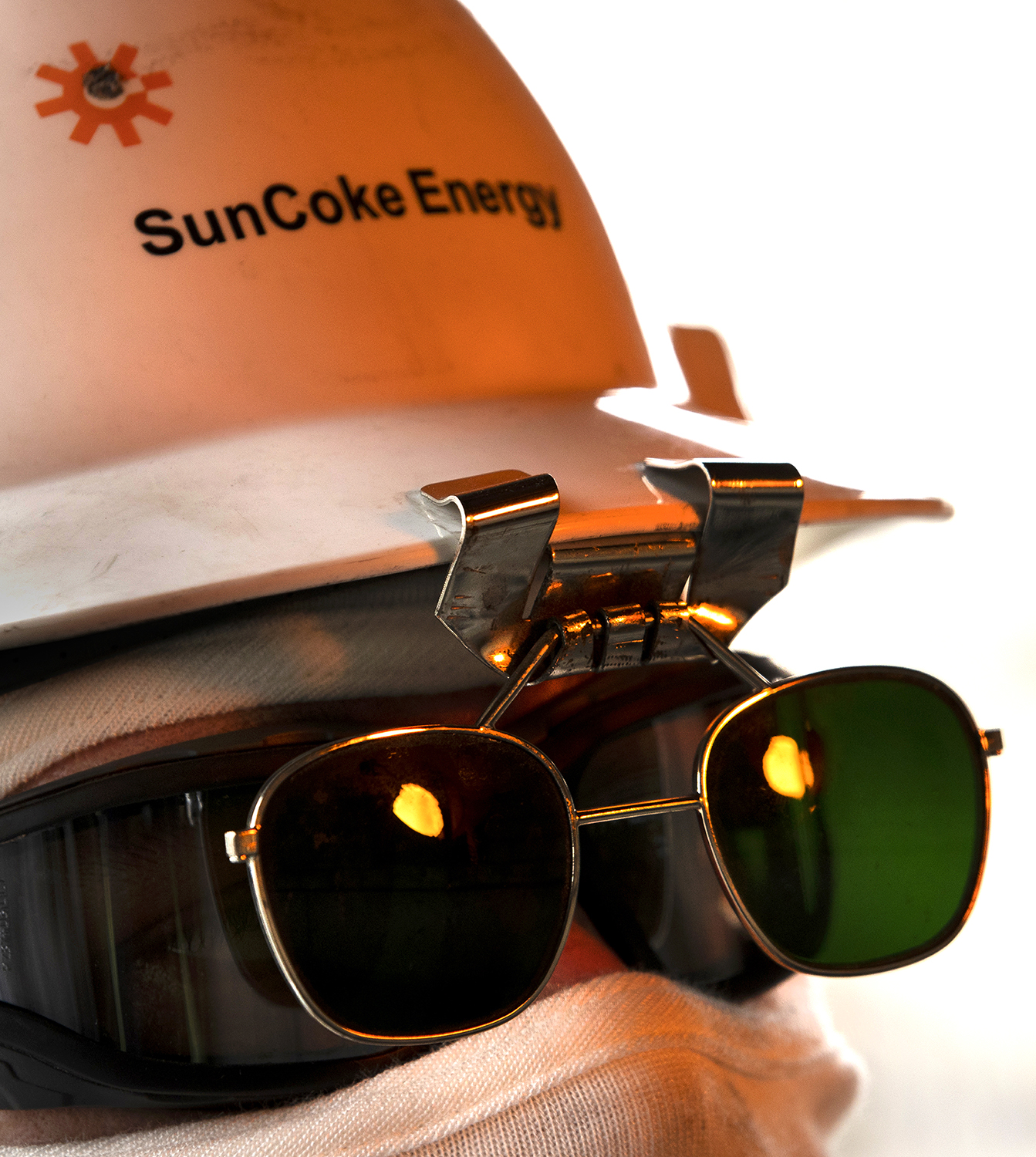 An employee wearing a hard hat that says SunCoke Energy is fully clad in protective gear. The image is a tight shot of the employee's face, which is not visible beneath the hat, dark safety glasses, and neck protector.