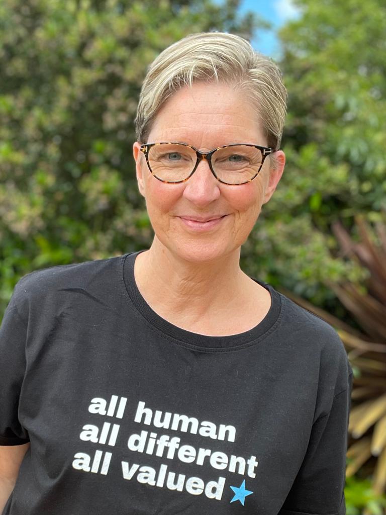 a lady called lesley harvey stood up in a garden wearing glasses and a black t shirt with the slogan, all human, all different, all valued