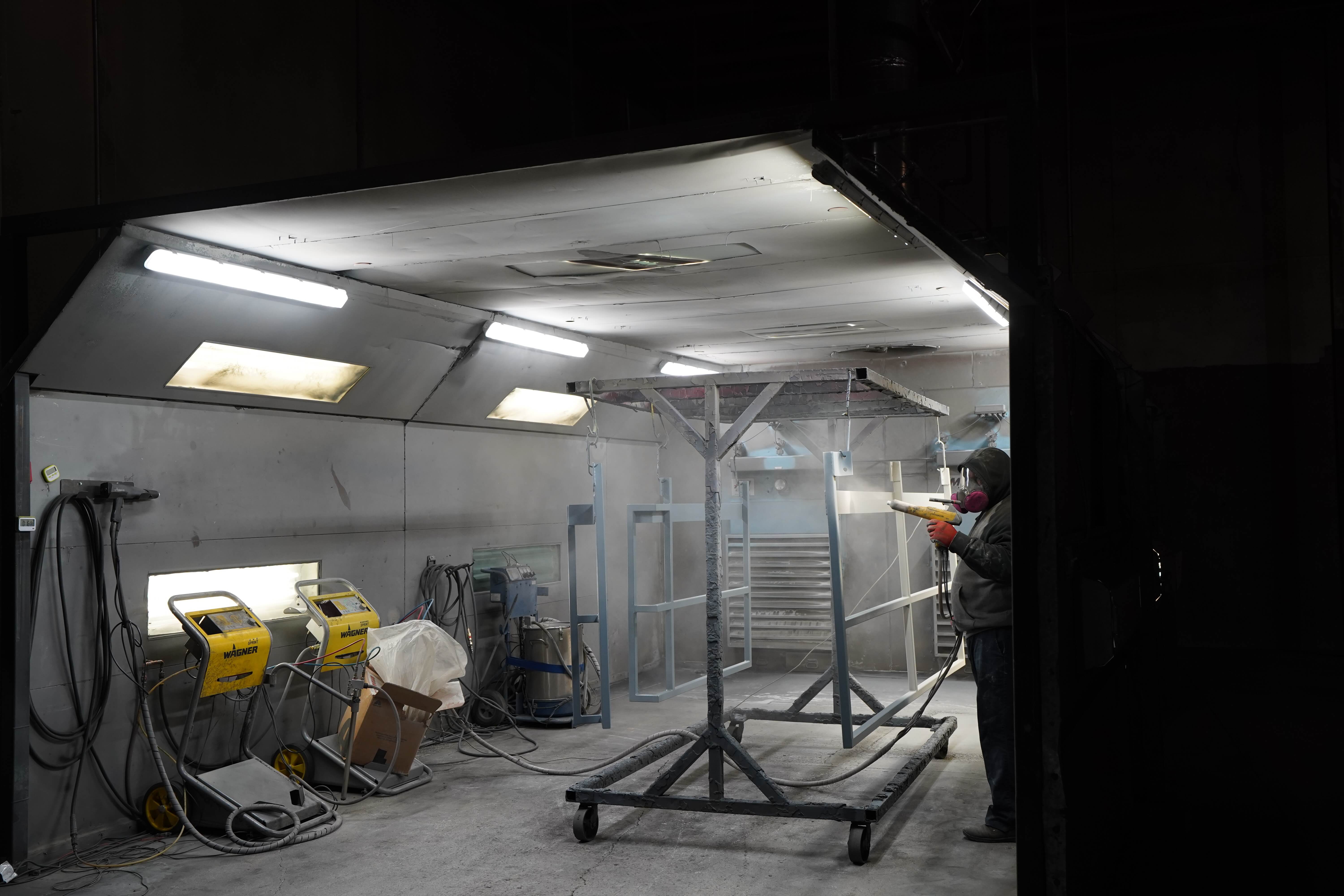 This image shows a powder coating spray booth or painting enclosure in an industrial workshop. On the right side, a worker wearing protective gear appears to be spraying or working with a powder coating gun. The booth has bright fluorescent lighting in the ceiling and white walls to maximize visibility. In the center of the booth is a metal rack or frame on wheels that appears to be used for hanging items during the coating process. On the left side, there are yellow powder coating machines or equipment carts with hoses. The environment is designed for safely applying powder coating to metal parts in a controlled space with proper ventilation and lighting. This spray booth is part of the powder coating process where fine particles are electrostatically applied to metal surfaces before being cured in an oven.