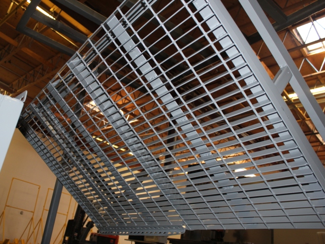 Industrial metal walkway or platform with galvanized steel grating, angled diagonally against a warehouse interior with warm-toned lighting and structural framework