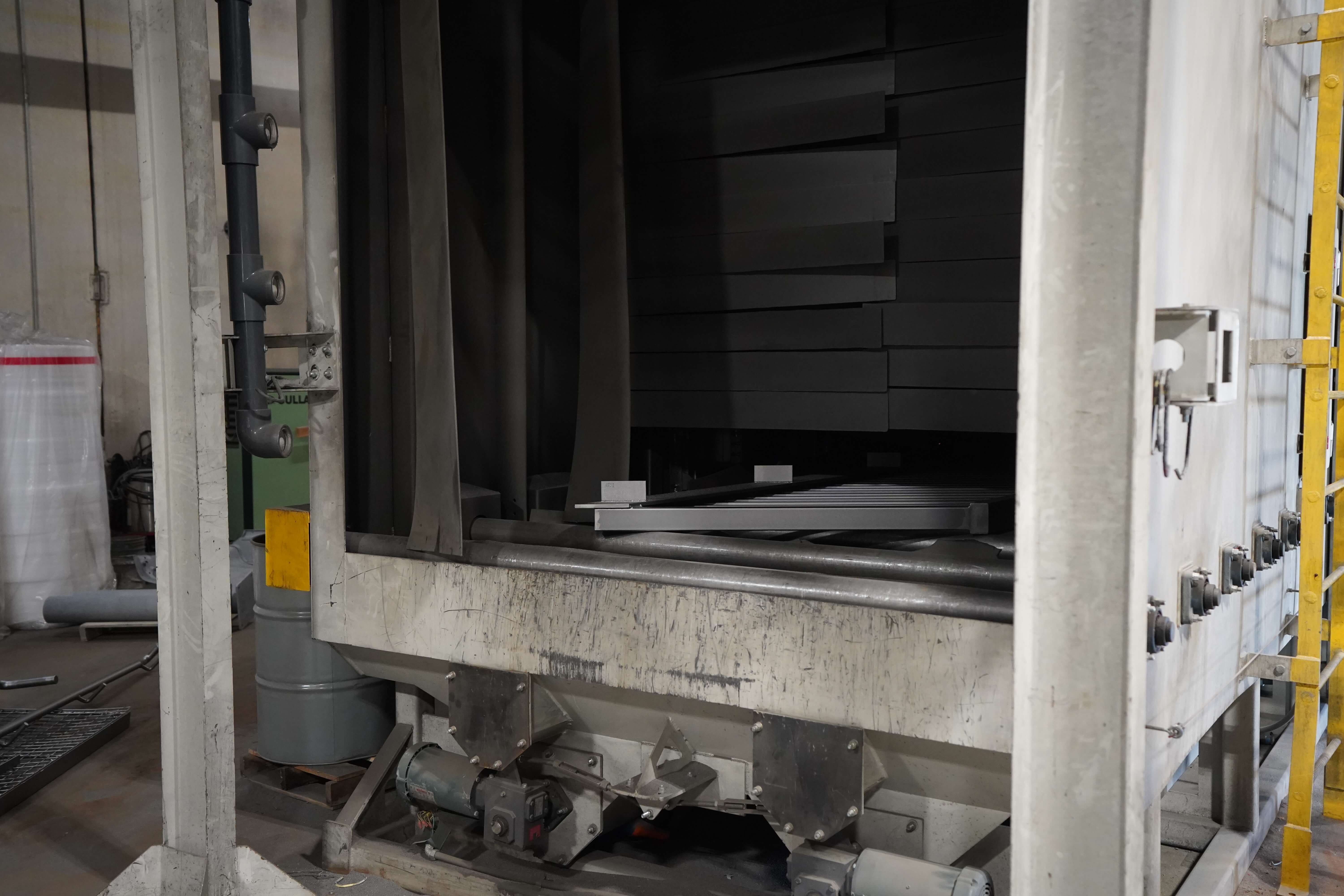 Industrial sand blasting machine used for metal surface preparation in a powder coating facility. The white cabinet structure has an internal chamber where abrasive media is propelled against metal parts to remove rust, old paint, and surface contaminants. Metal components can be seen inside the chamber, with conveyor or support mechanisms visible. The machine features control panels along the side, ventilation piping for dust collection, and robust structural elements. This equipment represents a critical pre-treatment stage where metal surfaces are cleaned and textured before powder coating application.