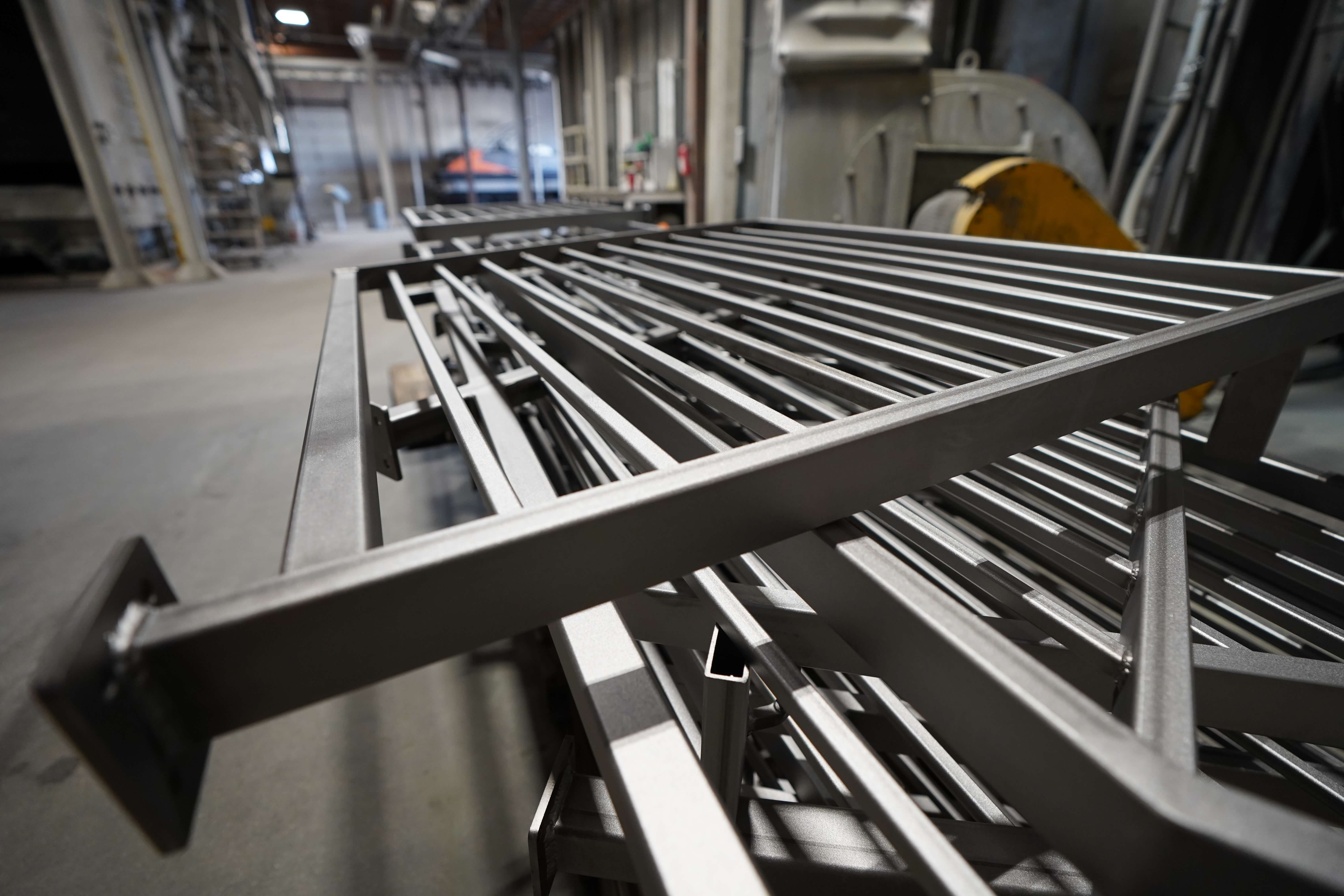 The image shows a close-up view of metal railings or fence panels in a fabrication workshop. In the foreground, there's a metal railing section with horizontal bars housed within a rectangular frame, all with what appears to be a black or dark powder-coated finish. Multiple similar panels can be seen stacked or arranged in the background