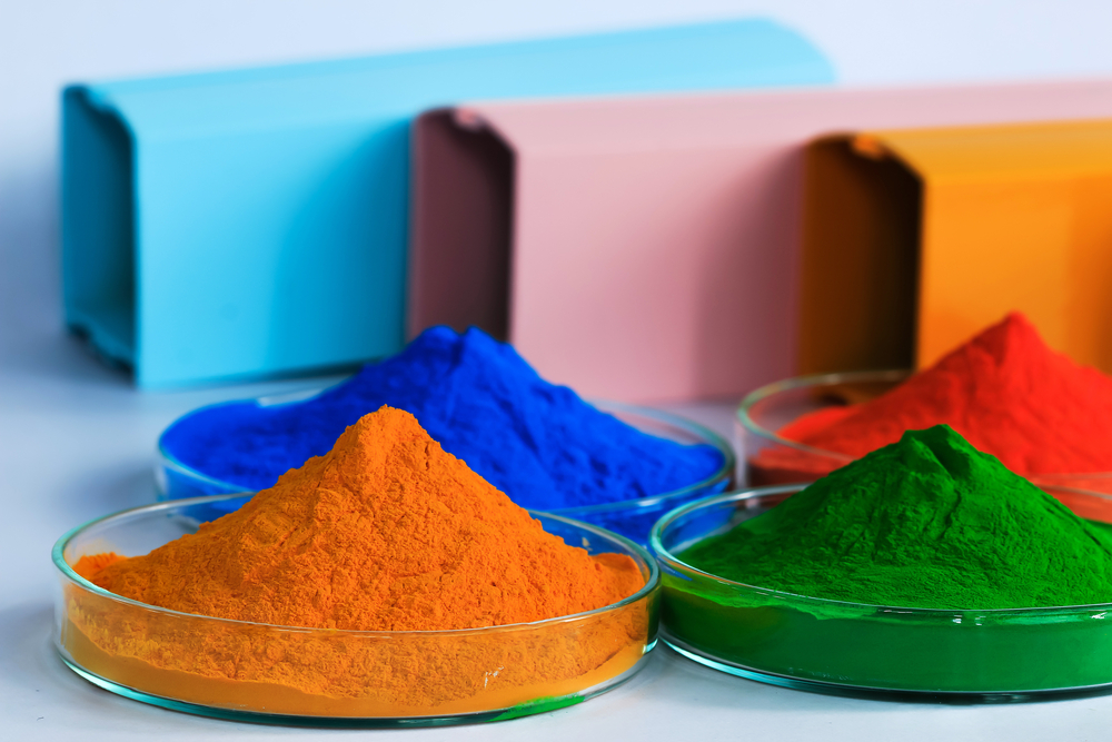 Vibrant powder coating pigments in orange, blue, green, and red, displayed in glass petri dishes with matching colored boxes in the background