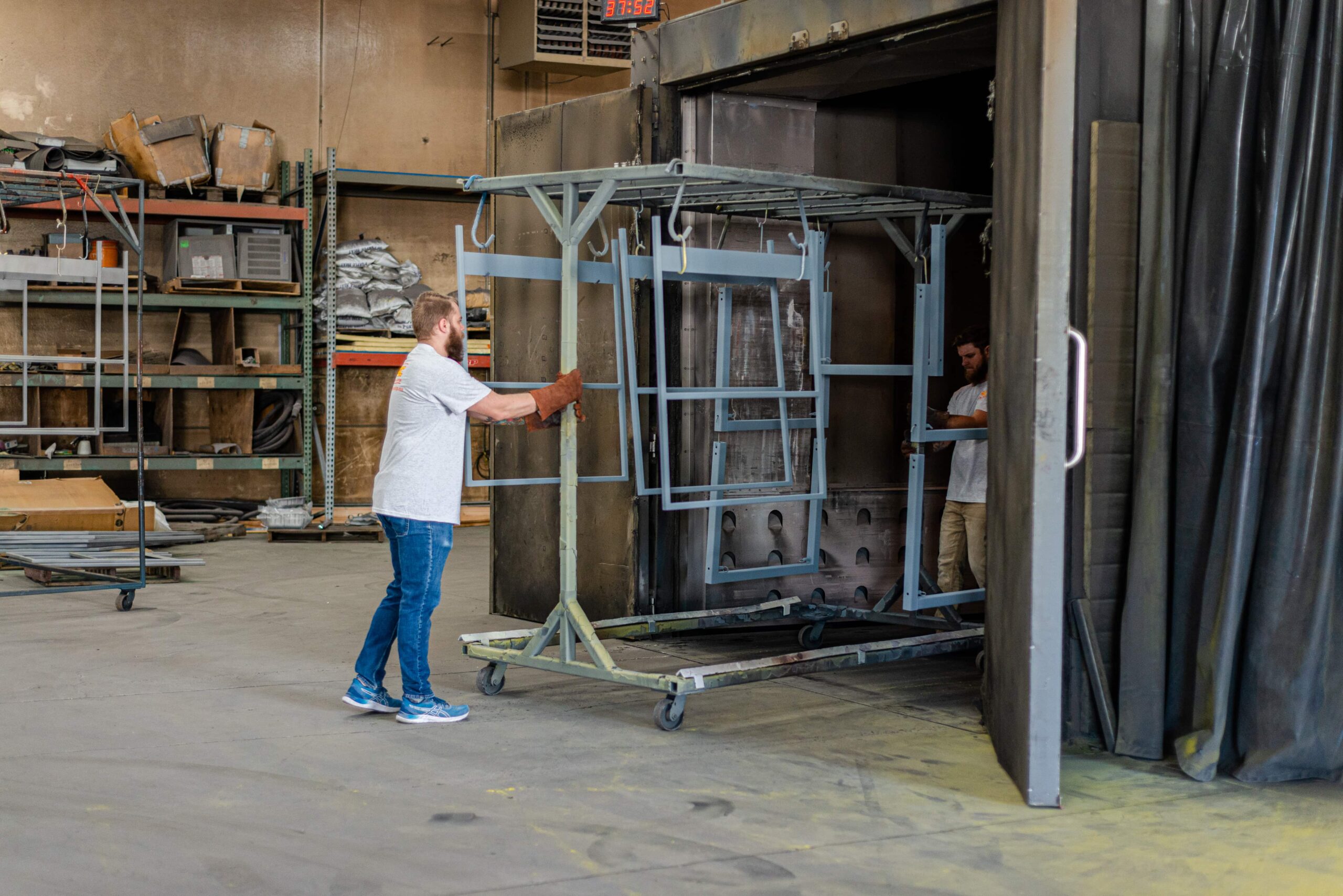Two workers in a powder coating workshop managing metal frames. One worker in a white shirt and jeans handles blue-painted metal frames on a wheeled rack cart, while another worker is visible inside a large industrial curing oven. The facility features storage shelving stocked with supplies, a digital display timer, and concrete flooring typical of metal finishing operations.