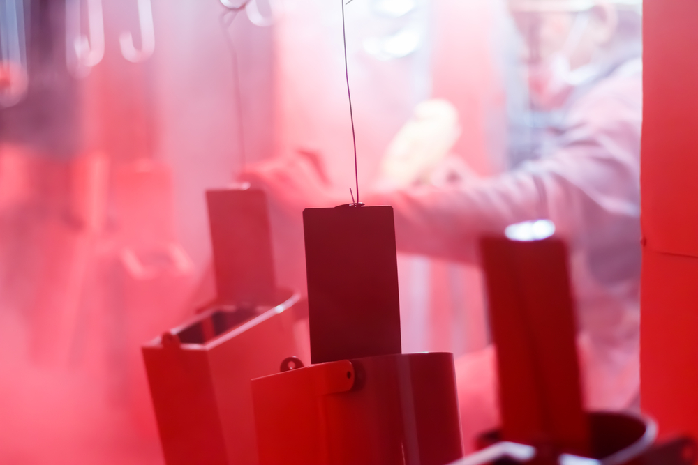 Close-up of powder coating equipment with rectangular metal blocks in a red-tinted industrial setting, showing hanging components in a powder coating preparation area