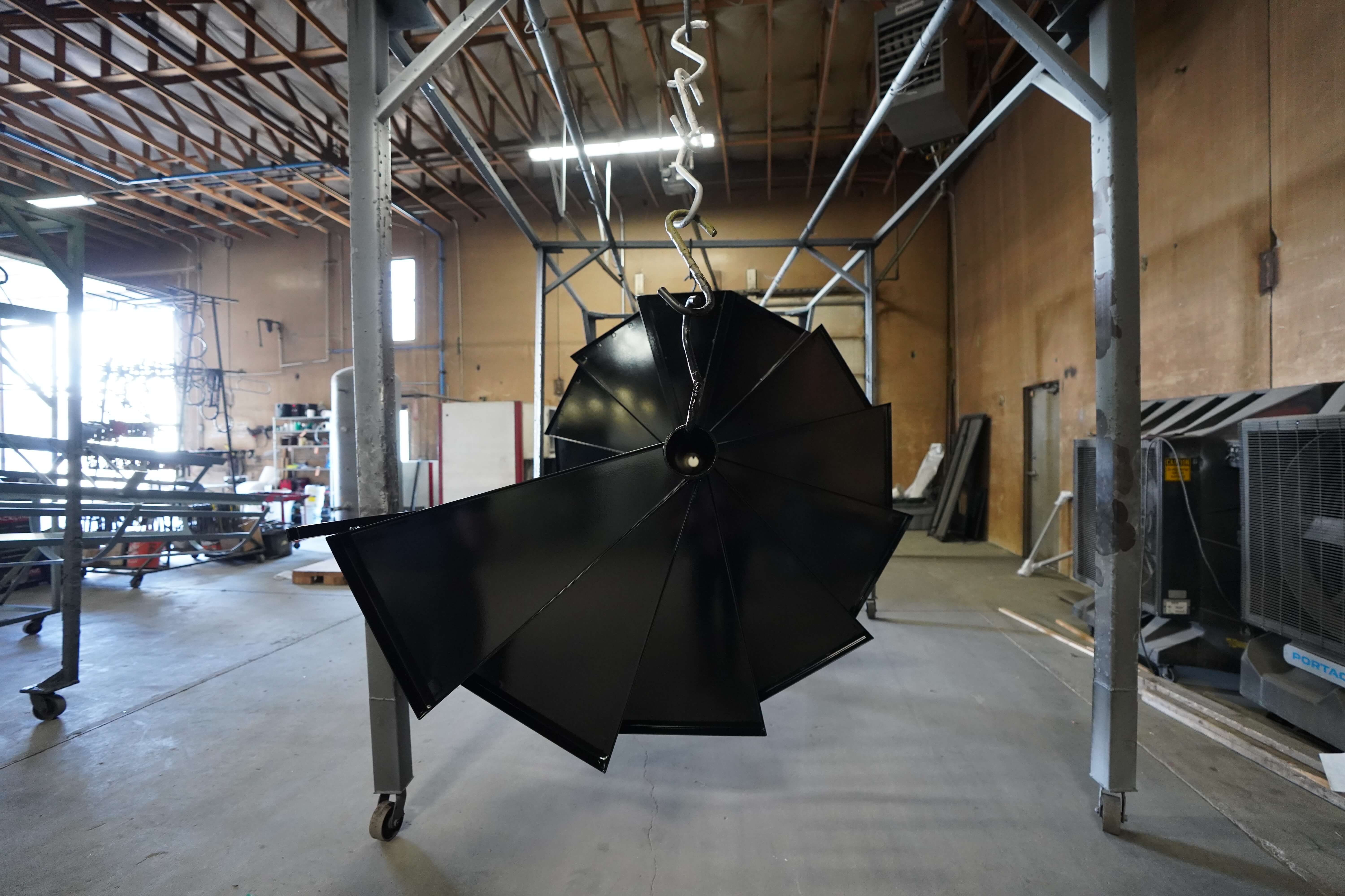 This image shows a black spiral staircase component suspended from hooks on a metal rack structure in what appears to be an industrial workshop. The spiral metal piece has been freshly powder coated or painted in a glossy black finish and is hanging to dry or cure. It's suspended from a wheeled metal frame with a rectangular shape, allowing it to be moved around the workshop.