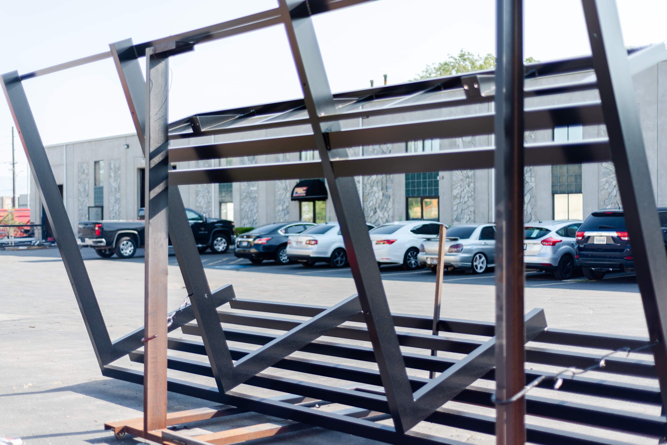 This image shows a powder-coated metal frame structure positioned in a business parking lot. The black metal framework appears to be a partially assembled sign or display structure that has been fabricated and finished with powder coating. Through the metal framework, several vehicles can be seen parked in front of what looks like an industrial or commercial building. The sturdy steel construction demonstrates the durability and professional finish that powder coating provides for outdoor metal structures. The image showcases both the structural components and the finished appearance of commercial metal fabrication work.