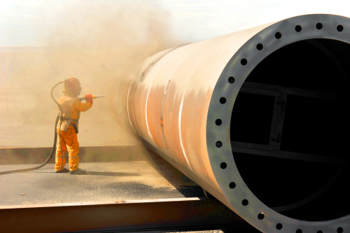 Worker in bright orange protective suit standing near a large industrial pipe with bolted flange, illuminated by soft ambient light