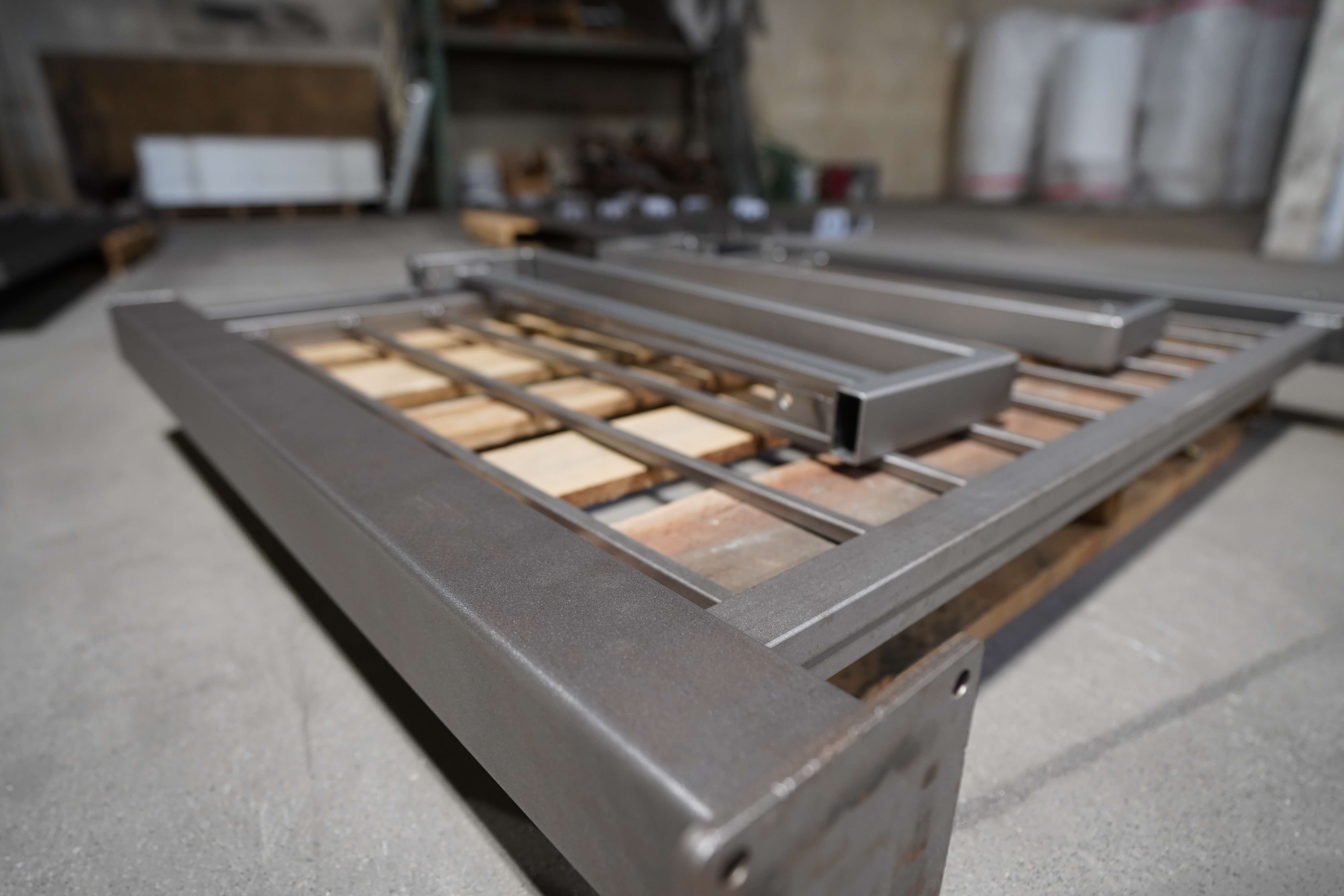 This image shows a powder-coated or painted metal gate frame lying on wooden pallets in what appears to be a workshop or fabrication facility. The frame has a silver/gray finish and consists of rectangular metal tubing assembled in a gate or door structure. The metal frame appears to be a finished product resting on wooden supports, likely after going through a coating process. The workshop environment has a concrete floor, with various supplies and equipment visible but blurred in the background. This appears to be part of a metal fabrication process where custom gates or security doors are manufactured and finished with protective coatings.
