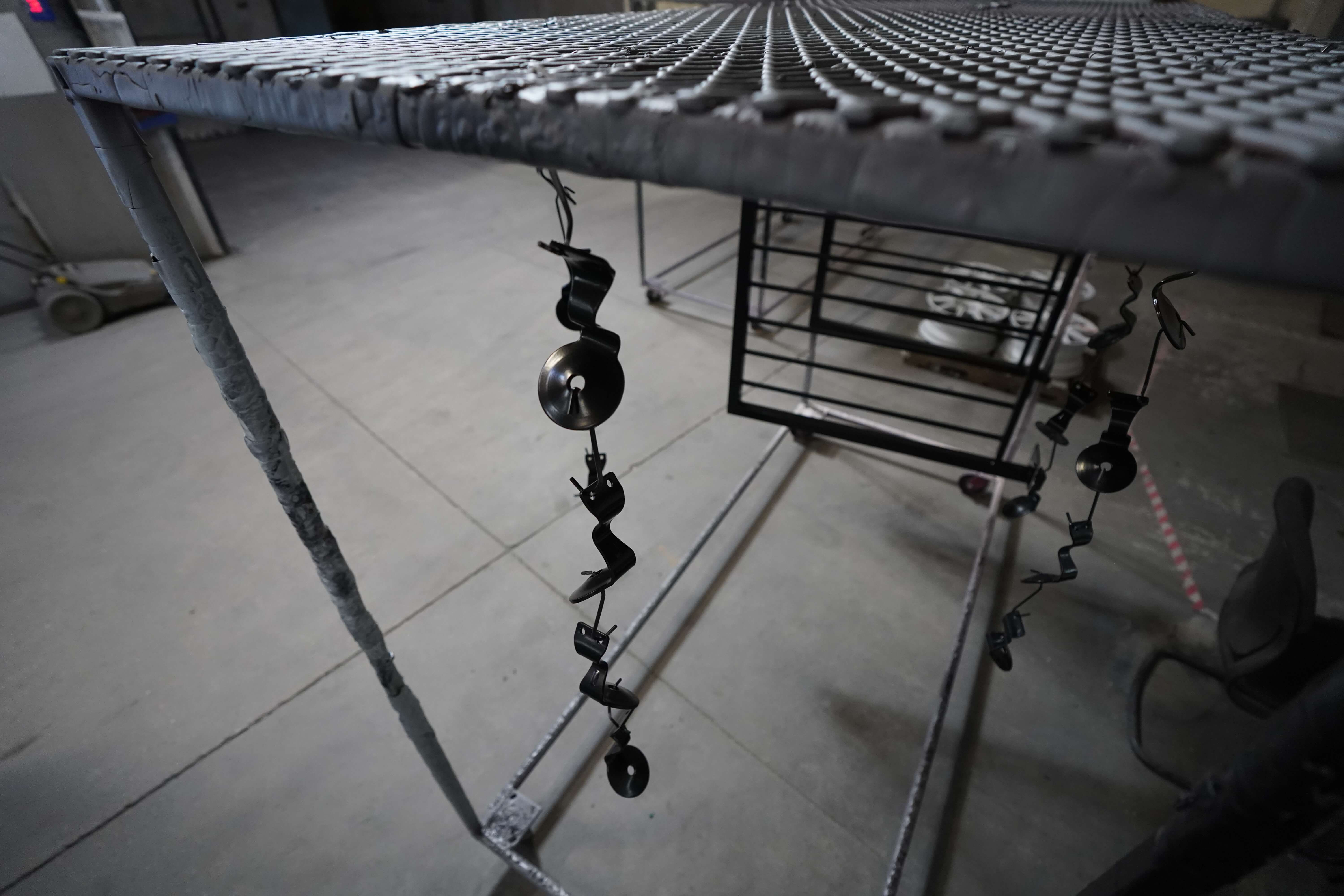 The image shows an industrial manufacturing setup with expanded metal mesh or grating visible from below. At the top of the image is a large sheet of expanded metal mesh with a diamond pattern stretching across the frame. Hanging from the mesh are several spiral or twisted metal elements with circular discs or pulleys attached, which appear to be part of a hanging or mounting system. In the background, you can see what looks like a black metal railing panel or gate similar to the one from the first image. The setting has a concrete floor and appears to be in the same industrial manufacturing facility seen in previous images, likely part of the metal fabrication process for railings or other custom metal products.