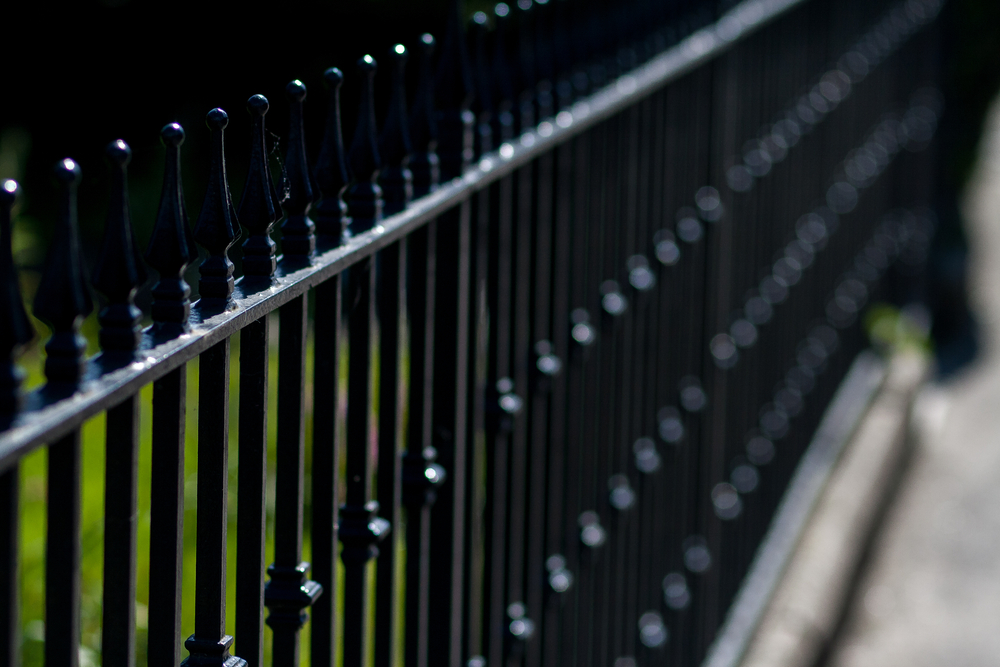 A close-up image of a black wrought iron fence extending into the distance with a shallow depth of field. The image captures the elegant detail of the vertical spindles with decorative finials along the top rail. The fence appears to be powder-coated in a glossy black finish that highlights its clean lines and ornamental elements. The background is blurred but shows hints of greenery, creating contrast with the dark metal. This type of durable, powder-coated metal fencing combines aesthetic appeal with long-lasting protection against corrosion and weathering.