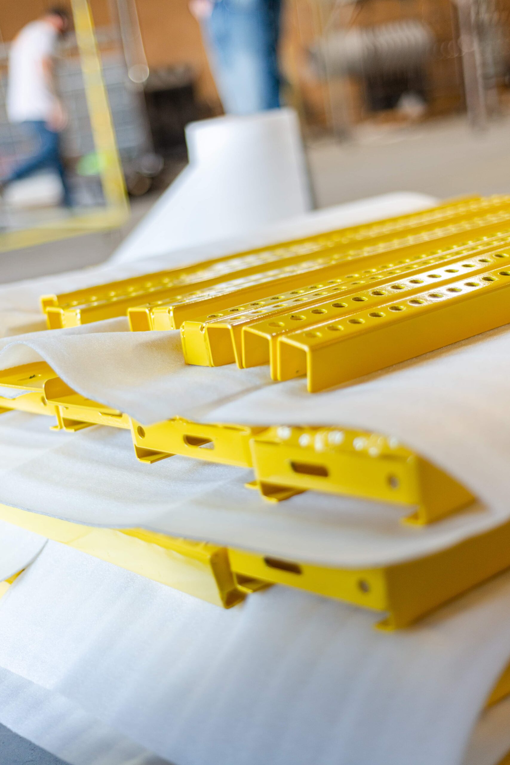 Bright yellow powder-coated metal components arranged on white protective paper in a manufacturing facility. The image shows various metal brackets and perforated channels with a glossy yellow finish, likely freshly coated pieces awaiting curing. A worker can be seen blurred in the background of the industrial workspace.