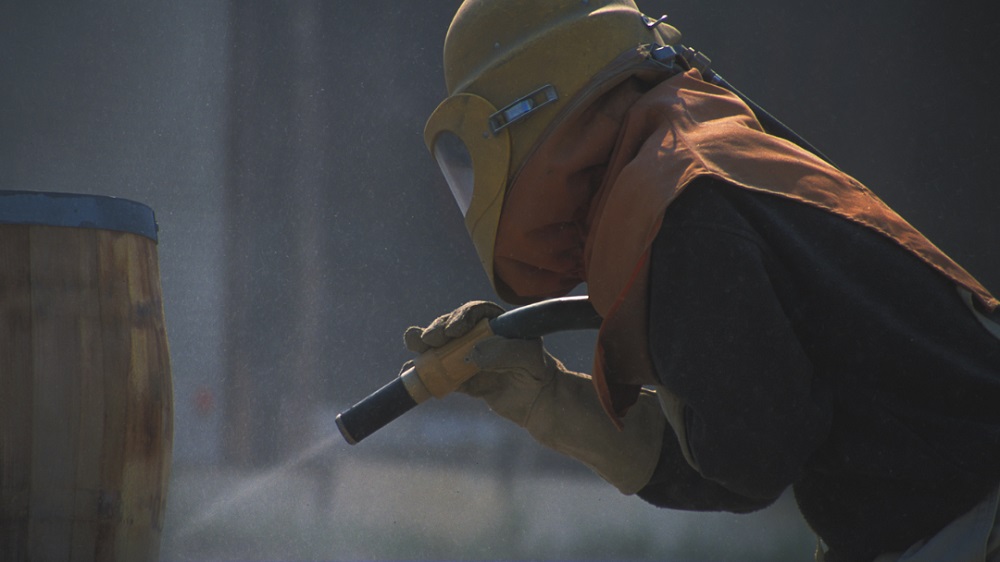Industrial worker in yellow hard hat and protective gear using a spray painting or sandblasting tool, surrounded by misty spray in a dimly lit industrial setting