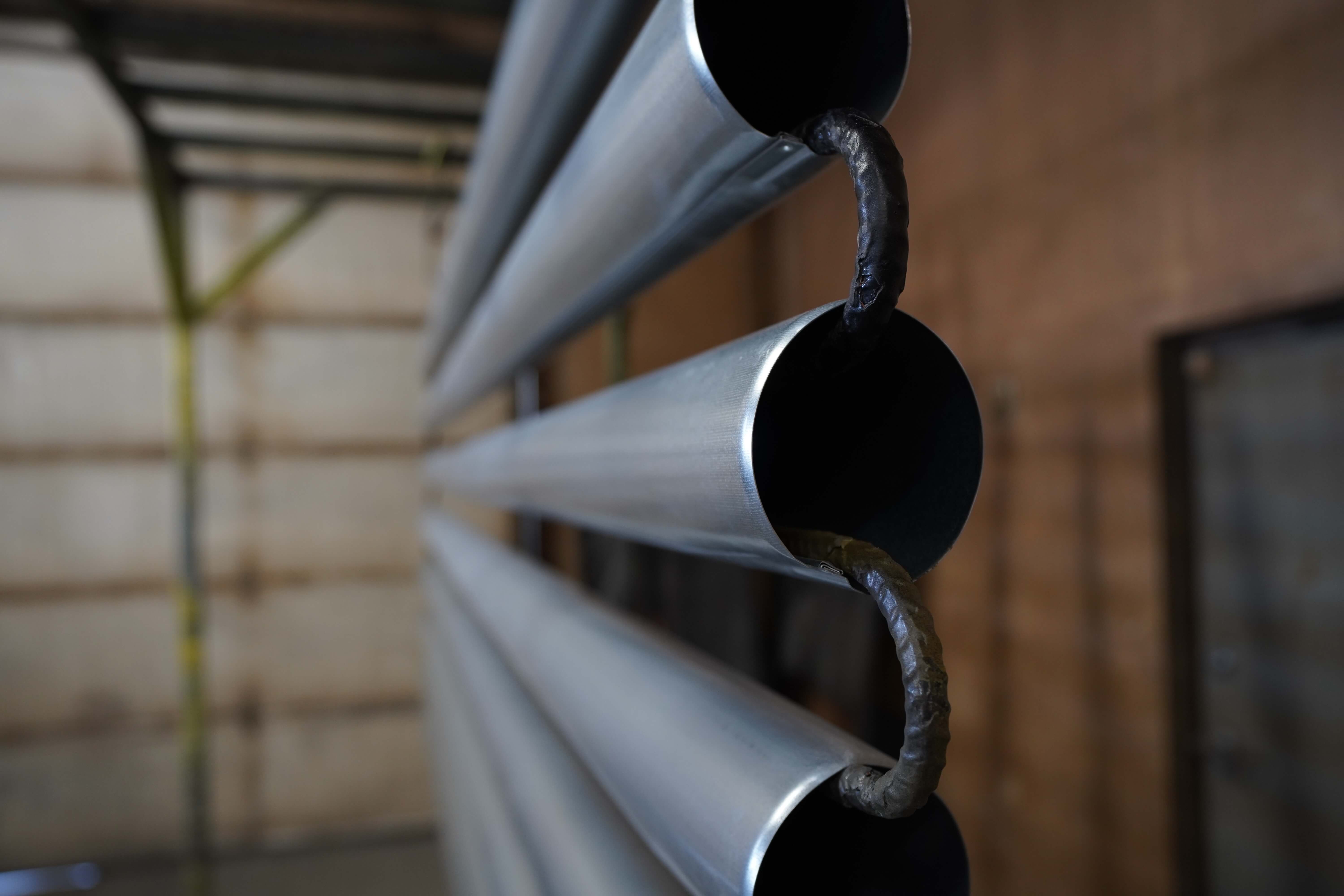 The image shows a close-up detail of what appears to be a modern metal railing or architectural element. Three horizontal brushed stainless steel or aluminum tubes are arranged in a parallel configuration, with custom-fabricated metal connectors joining them. The connectors have a handcrafted appearance with visible welding marks that connect each tube to the next. The perspective shows the tubes extending horizontally with the open hollow ends visible. The background shows what appears to be the same industrial manufacturing facility seen in previous images, with soft-focus wall panels. This appears to be a detail shot of a finished or nearly finished product from the metal fabrication workshop shown in the earlier images.
