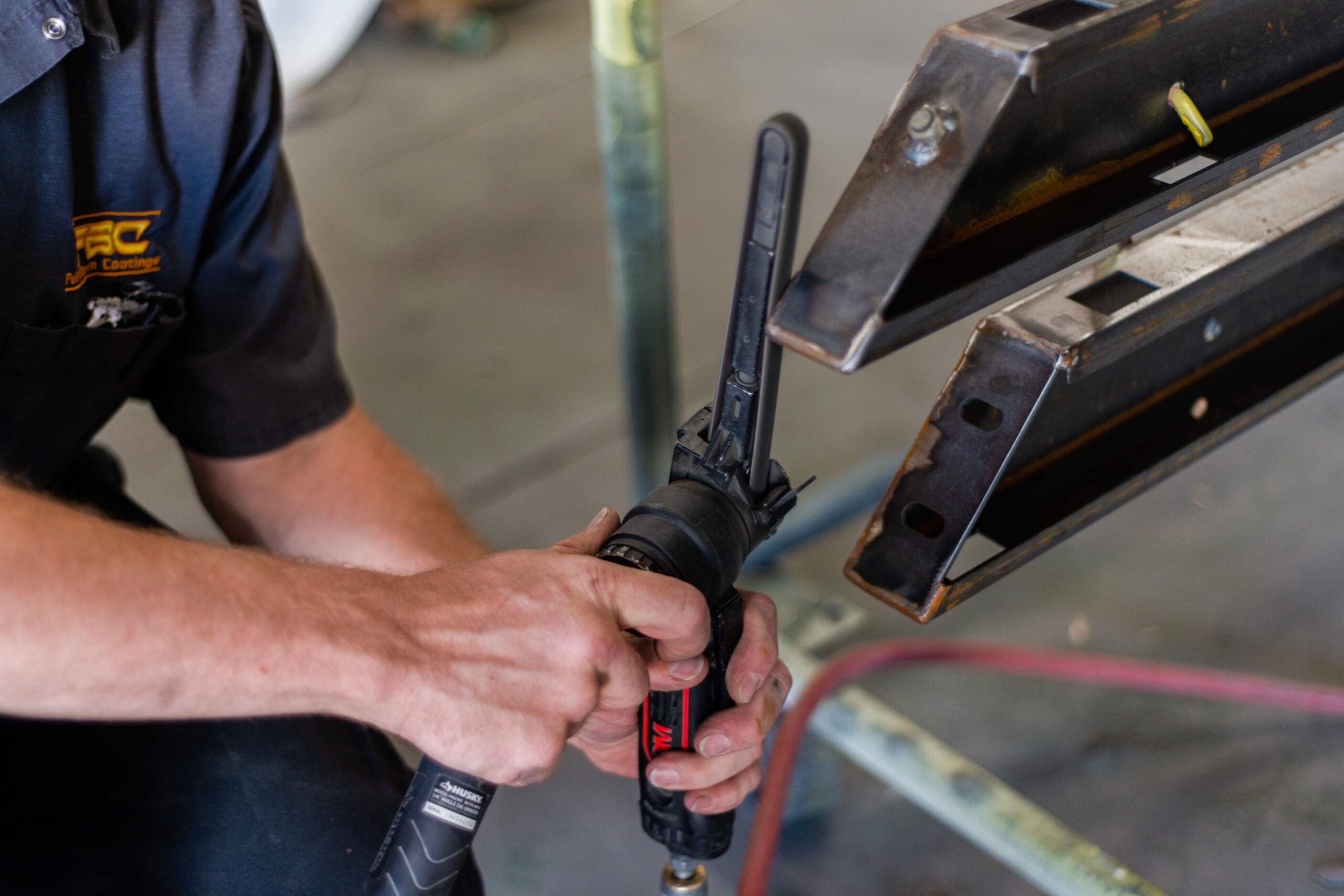 A close-up image of a technician working with metal components at a powder coating facility. The worker, wearing a dark uniform shirt with a company logo, is using a pneumatic or power tool to adjust or prepare metal brackets or frames. The technician's hands are clearly visible gripping the tool as they work on what appears to be metal structural components that will likely undergo powder coating treatment. The image captures the precision and hands-on craftsmanship involved in metal preparation prior to the coating process.