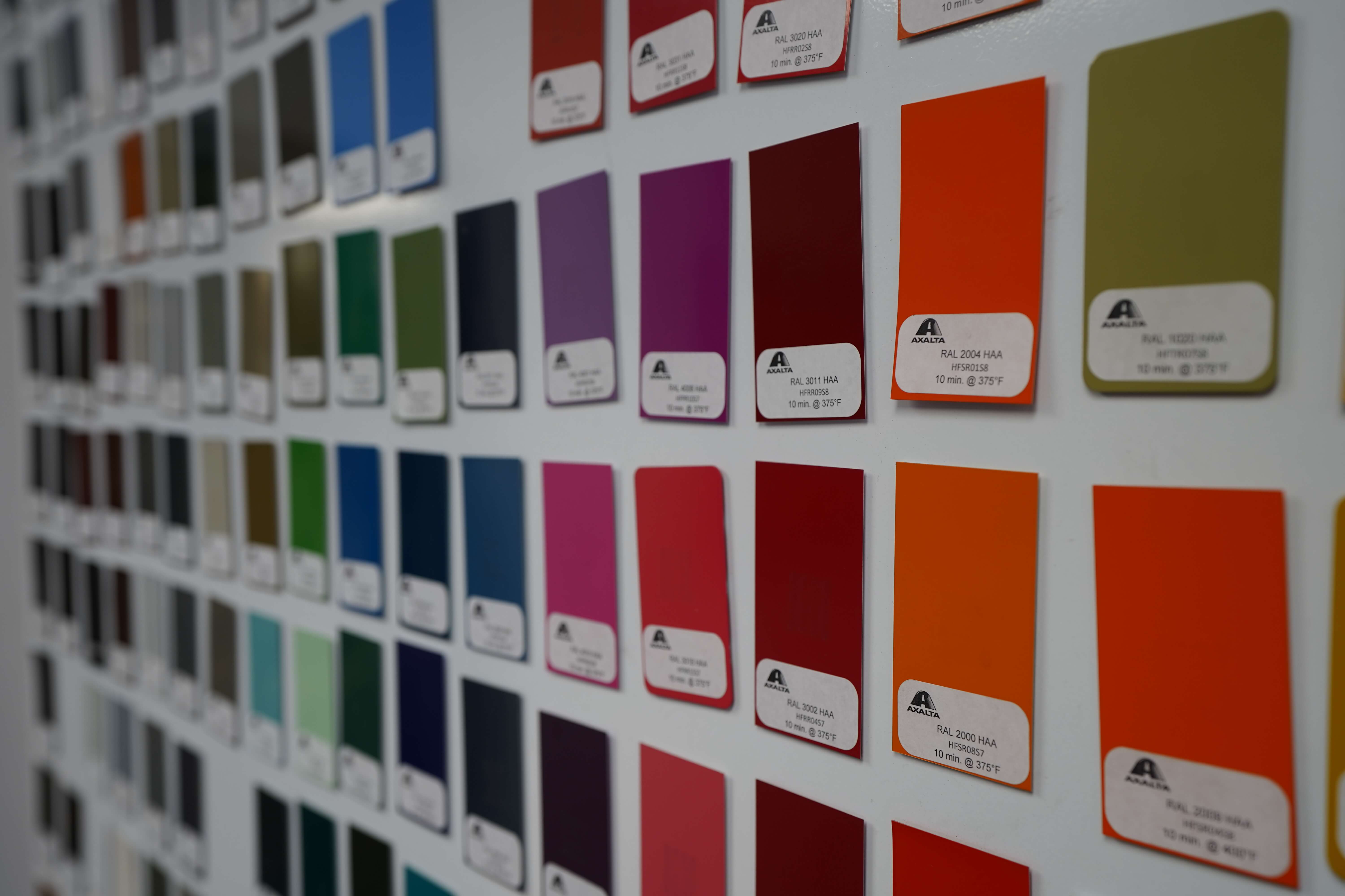 Close-up of a powder coating color sample display board showing a variety of vibrant finish options. The board features neatly arranged rectangular metal samples with a wide spectrum of colors including purples, reds, oranges, blues, greens, and neutrals. Each color sample includes a small identification label with color code information and curing specifications. This comprehensive display showcases the range of powder coating finishes available for metal surface treatments, allowing customers to select their preferred color and finish type.