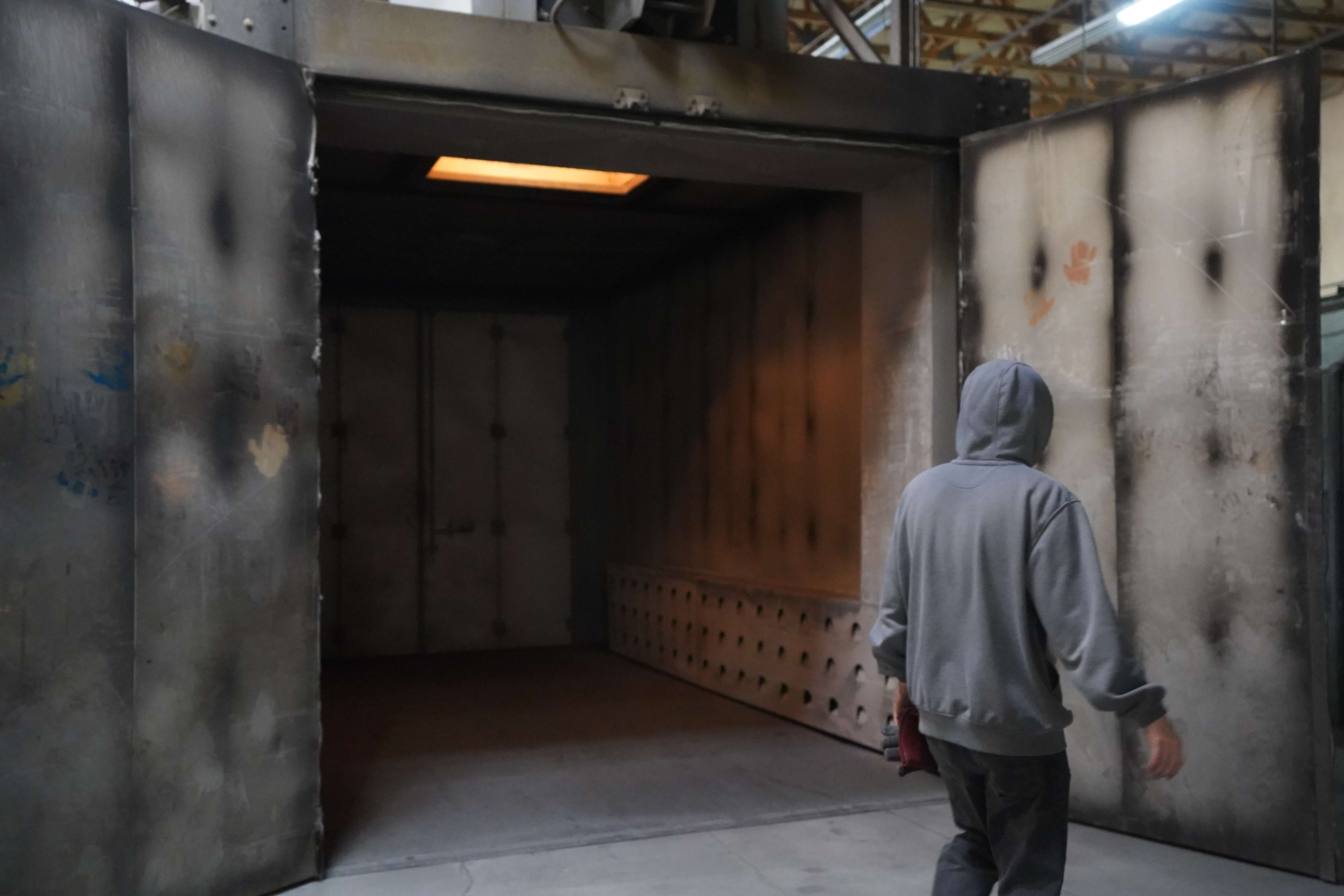 The image shows an industrial manufacturing or finishing area with a large open chamber that appears to be a paint booth or powder coating oven. In the foreground, a person wearing a gray hoodie is walking toward or entering the chamber, shown from behind. The chamber has metal walls that show signs of use with some smudges and markings. There's an orange/amber light visible at the top of the chamber, suggesting it might be a heating or curing element. The floor is concrete, and what looks like a ventilation or collection system can be seen at the bottom of the chamber. The industrial setting has a high ceiling with some structural elements visible above. This appears to be part of a metal fabrication or finishing process, possibly for the metal components shown in previous images.