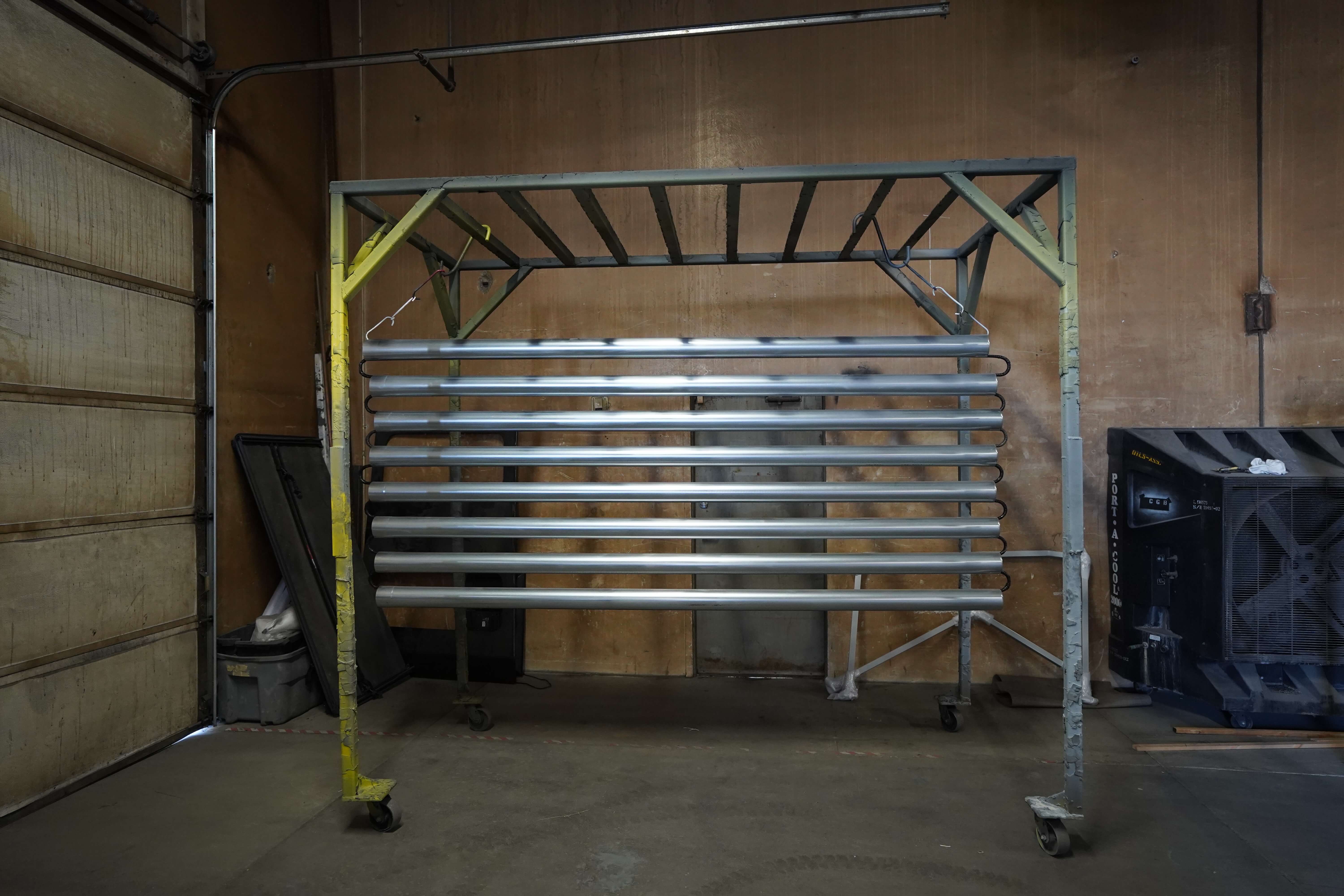 This image shows a metal rack system used for holding and displaying metal tubes or pipes. The rack consists of a wheeled metal frame structure with silver-colored aluminum tubes arranged horizontally across it. The frame is painted gray with some yellow markings visible on the left support. The structure is positioned against a wooden wall in what appears to be an industrial workshop or garage space, with a concrete floor. On the right side, there appears to be some equipment or packaging. This rack system likely serves as a drying station or storage solution for metal components after fabrication, coating, or other industrial processing. The garage door track is visible at the top of the image, indicating this is in a workshop with vehicle access.