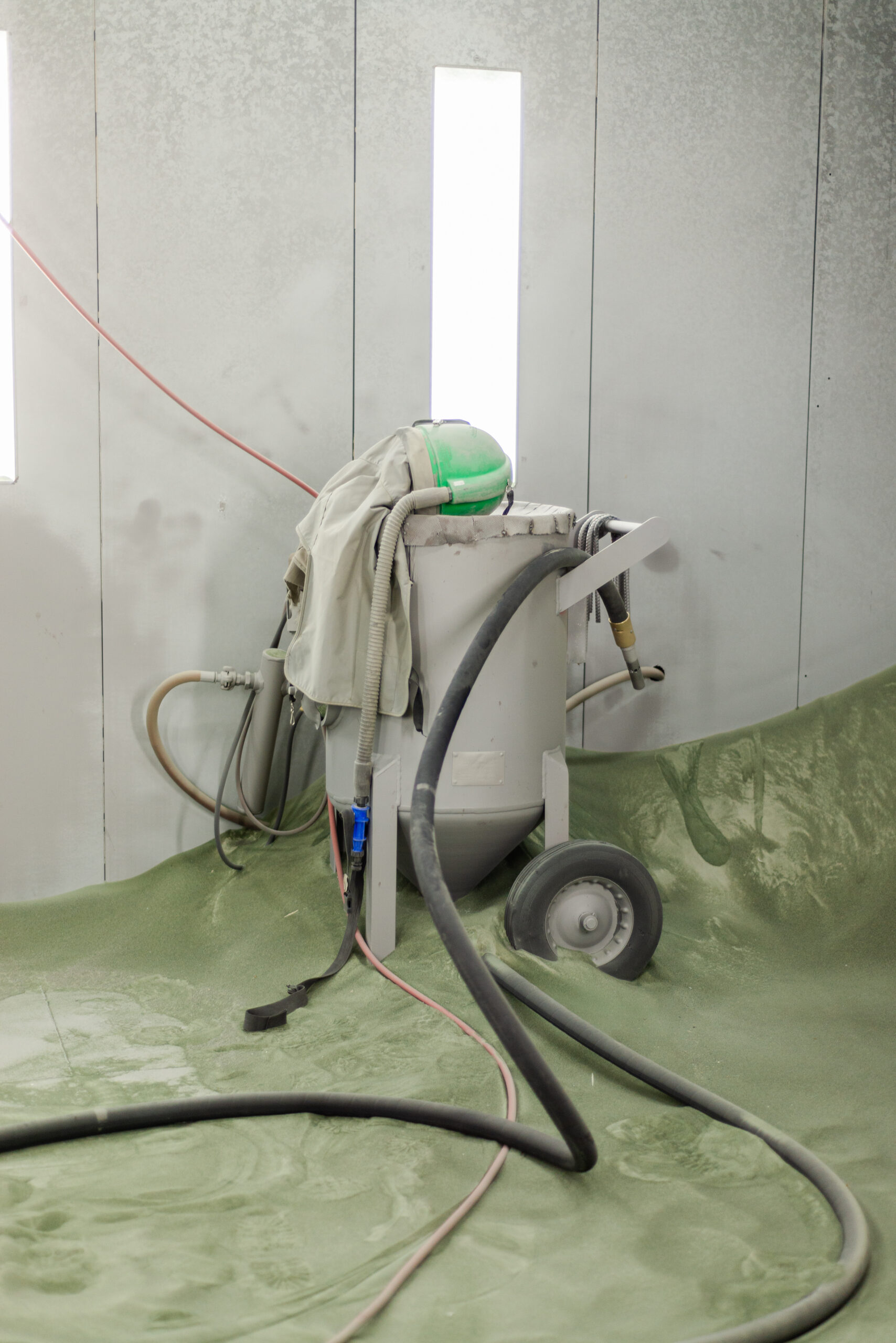 Industrial sandblasting equipment on wheels with attached hoses and green safety helmet, positioned in a spray booth with protective green floor covering for powder coating and metal resurfacing applications