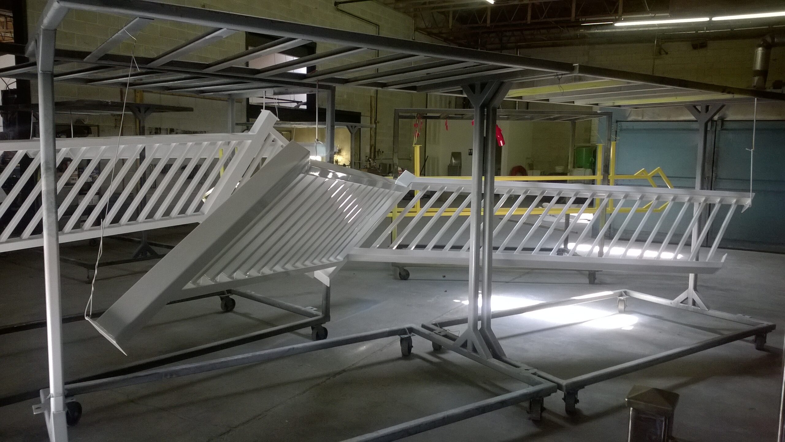 Industrial workspace with multiple white metal racks on wheels, featuring slatted designs, positioned under overhead structural framing in a large manufacturing facility with concrete floors and industrial lighting