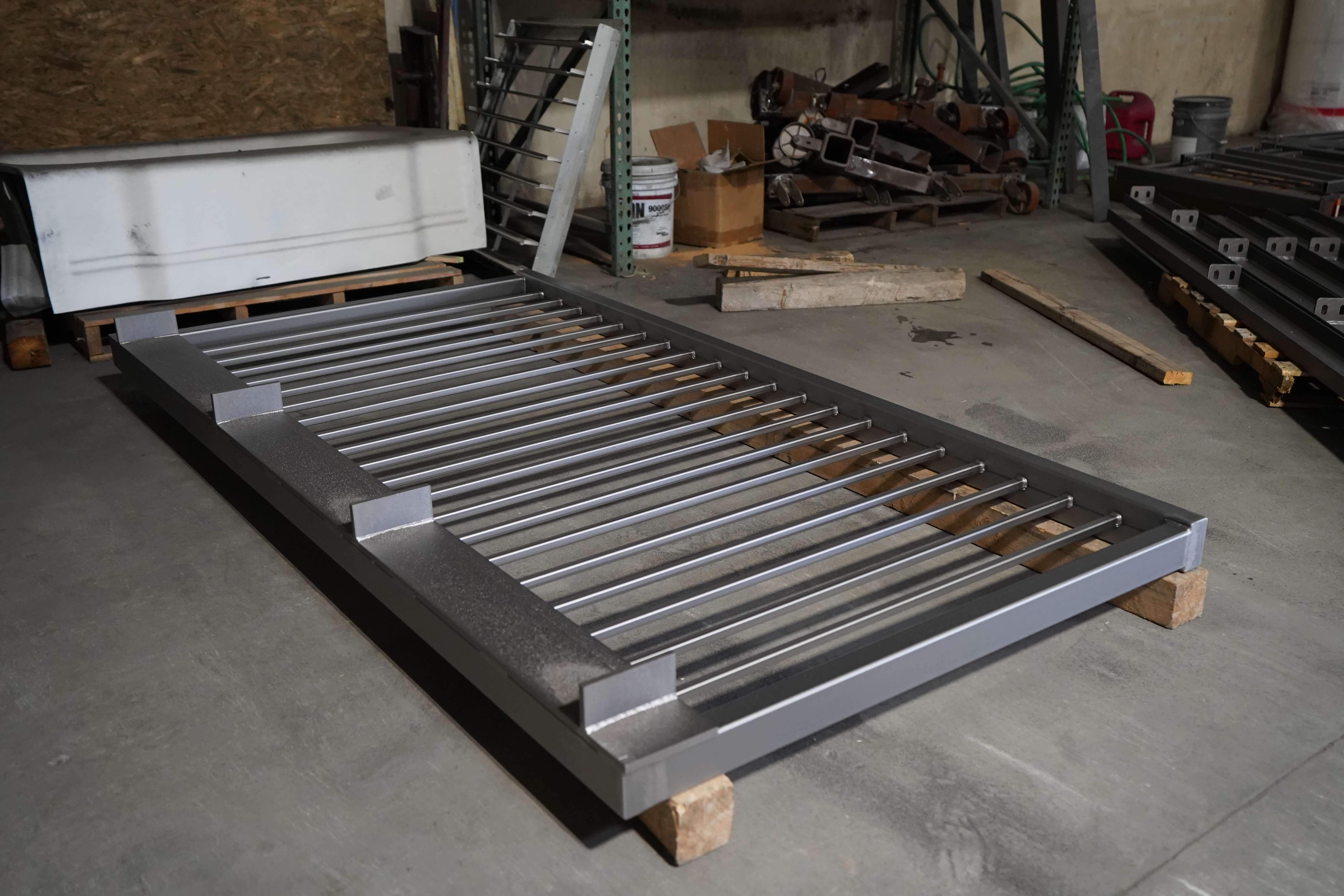 A finished powder-coated metal gate or railing panel resting on wooden blocks on a concrete floor in a manufacturing facility. The panel features a dark gray or textured finish on the frame with parallel silver/aluminum bars running horizontally across it. In the background are industrial storage racks containing various metal parts, supplies, and equipment used in the powder coating process. This demonstrates a completed product from the metal surface treatment company.