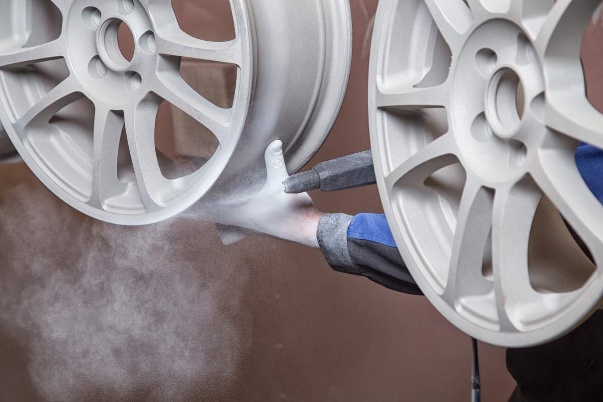 This image shows a powder coating process in progress. White aluminum car wheels or rims are being coated with a spray gun that is emitting a fine mist of white powder coating material. The spray gun, held by someone wearing blue and gray gloves, is directing the powder toward the wheel surface. The image captures the cloud of powder particles suspended in the air between the spray nozzle and the wheel surface. This process uses electrostatic charging to make the powder particles adhere to the metal surface before the coated wheels are cured in an oven, creating a durable and attractive finish. The wheels appear to have a multi-spoke design and are positioned for efficient coating of their visible surfaces.