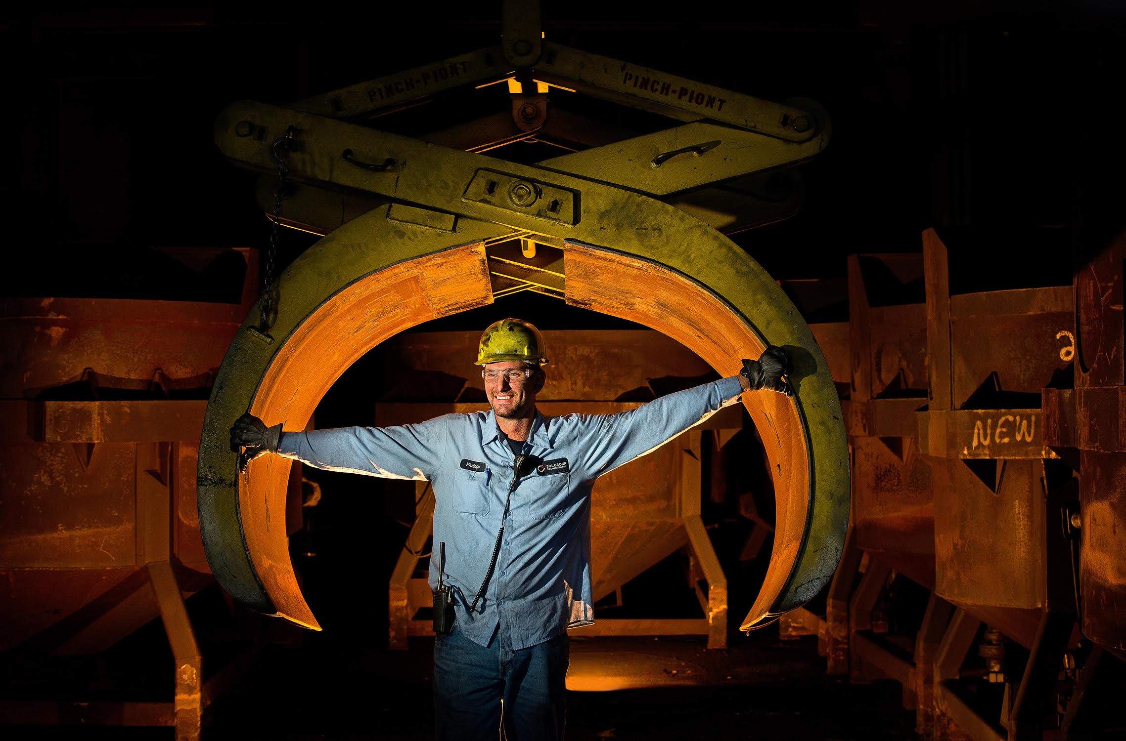 A male worker in a heavy industrial plant poses to have his photo taken. He is standing in front of a giant claw-type tool and has his arms stretched out, which helps show the size of the jaw. This is an environmental portrait of the worker, and uses special lighting to highlight the area around where he works. The worker is wearing proper safety equipment.